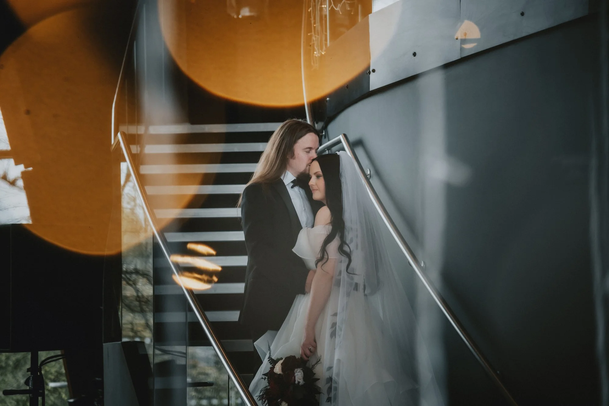 A couple in wedding attire sharing a tender moment on a staircase, with the man in a tuxedo and the woman in a wedding dress holding a bouquet of flowers.