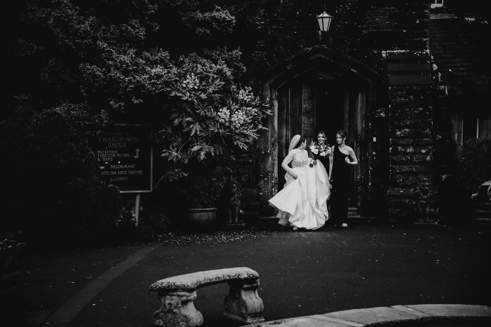 Black and white photo of three women, two in wedding dresses and one in a black dress, standing beneath a large wooden archway at a castle entrance, with a stone bench in the foreground and lush trees and a sign in the background.