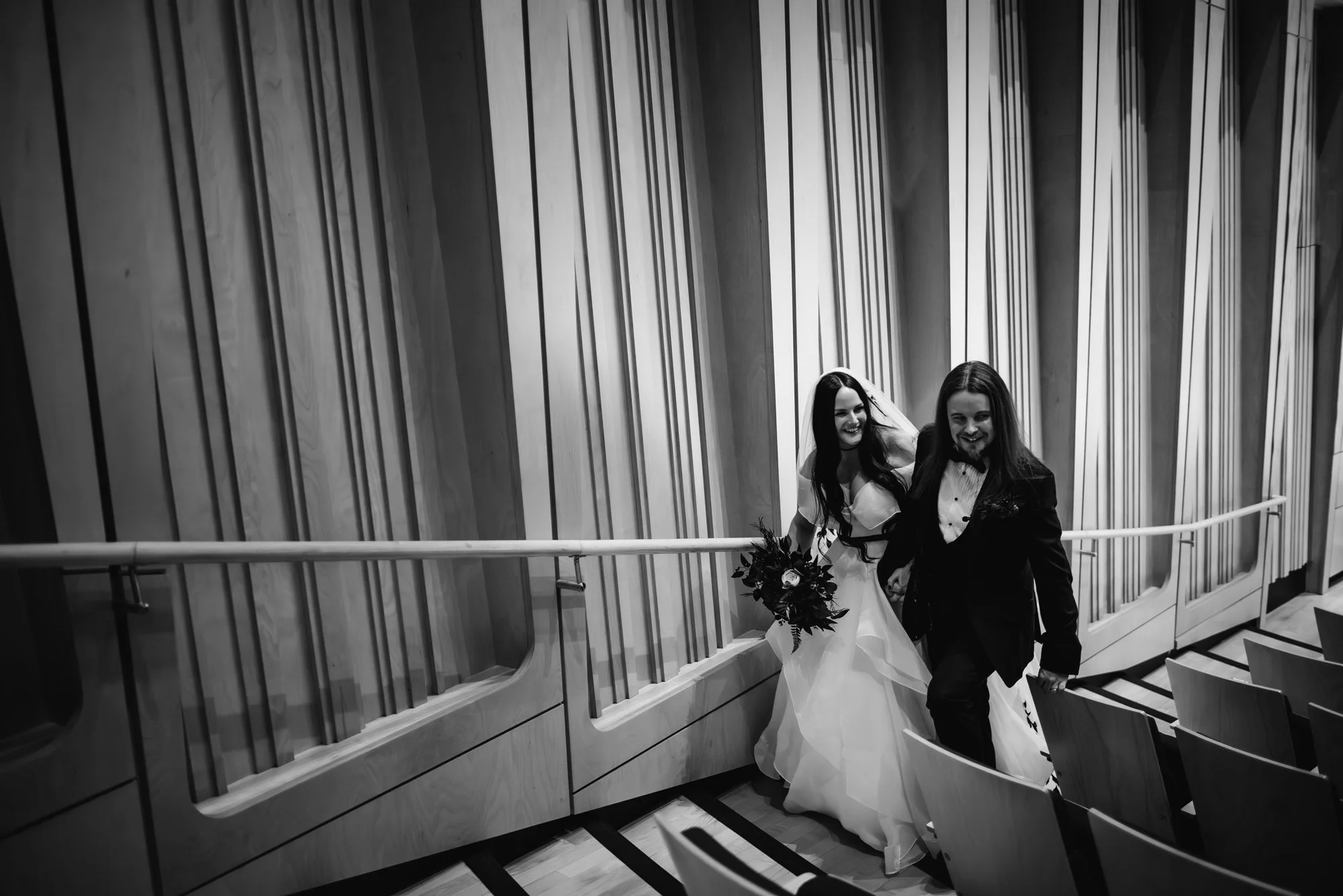 Black and white photo of a bride in a wedding dress and a groom in a tuxedo walking down a hallway with wooden paneling, smiling, and holding hands.