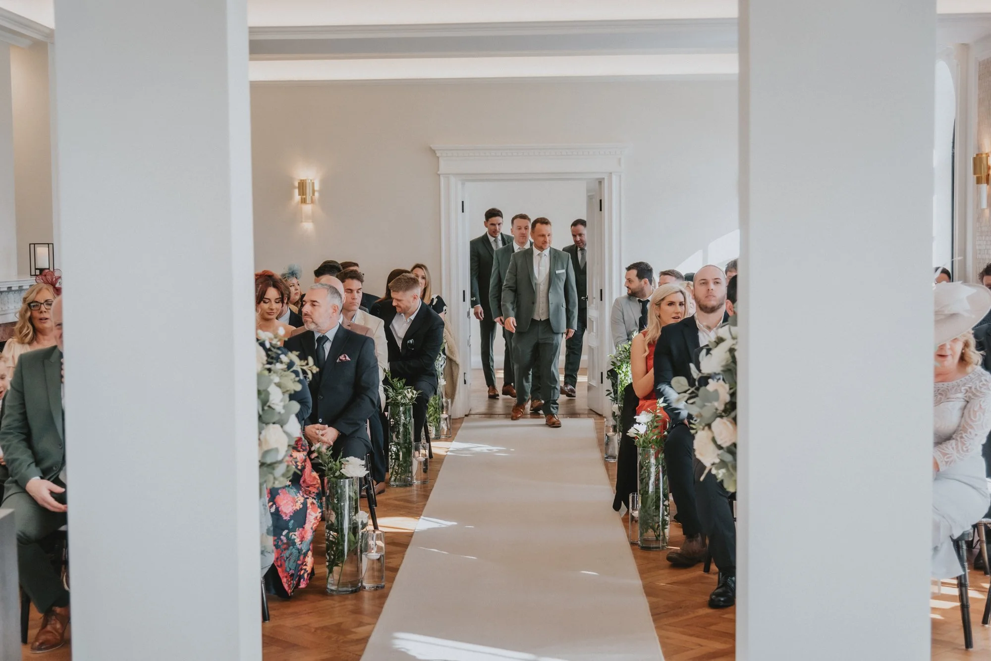 Guests seated in a wedding ceremony hall, with men in suits and women in dresses, some wearing hats, looking towards the aisle where a group of men in suits are walking in.