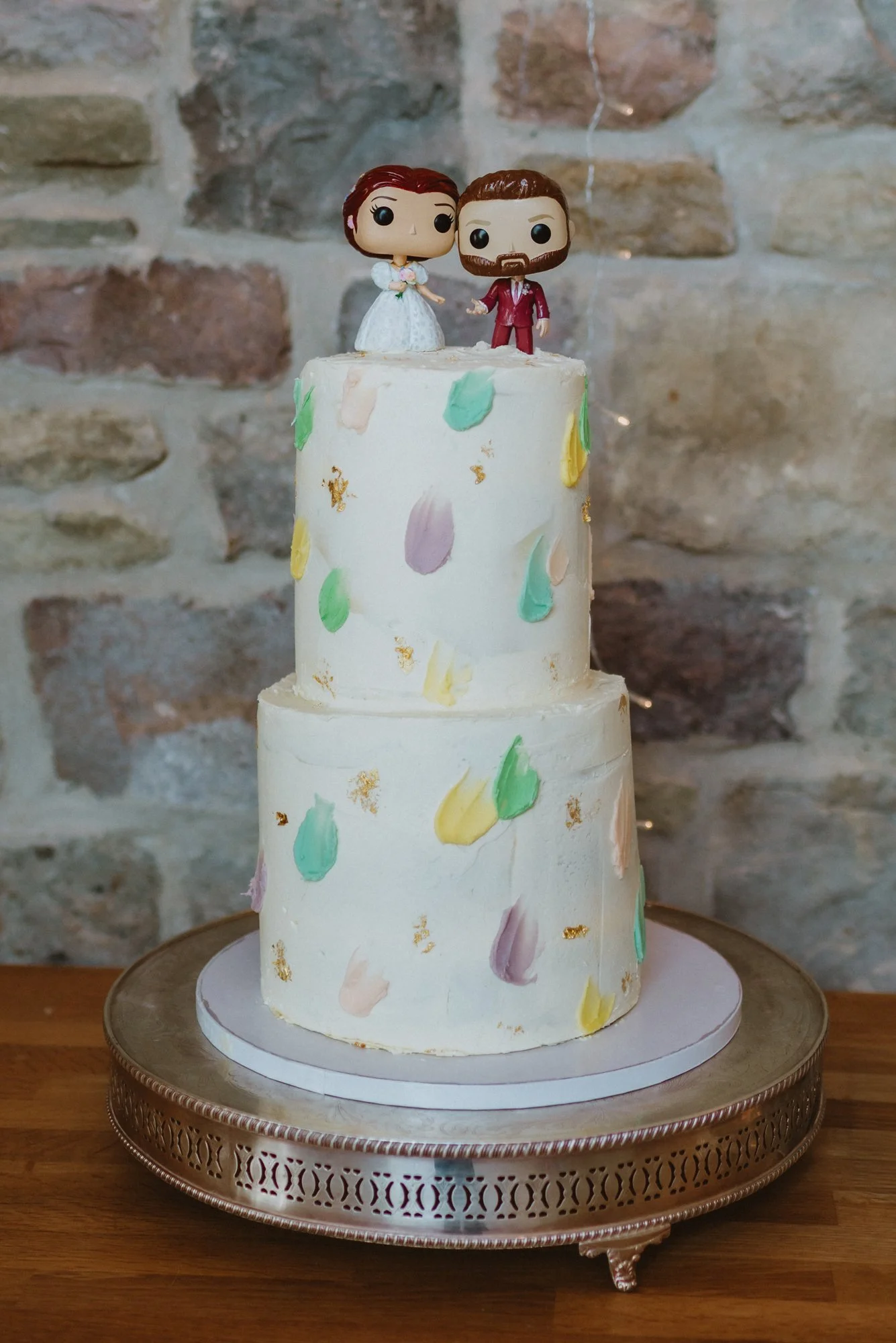 A two-tier wedding cake with pastel-colored abstract decorations, topped with a bride and groom toy figurines, on a decorative silver cake stand.