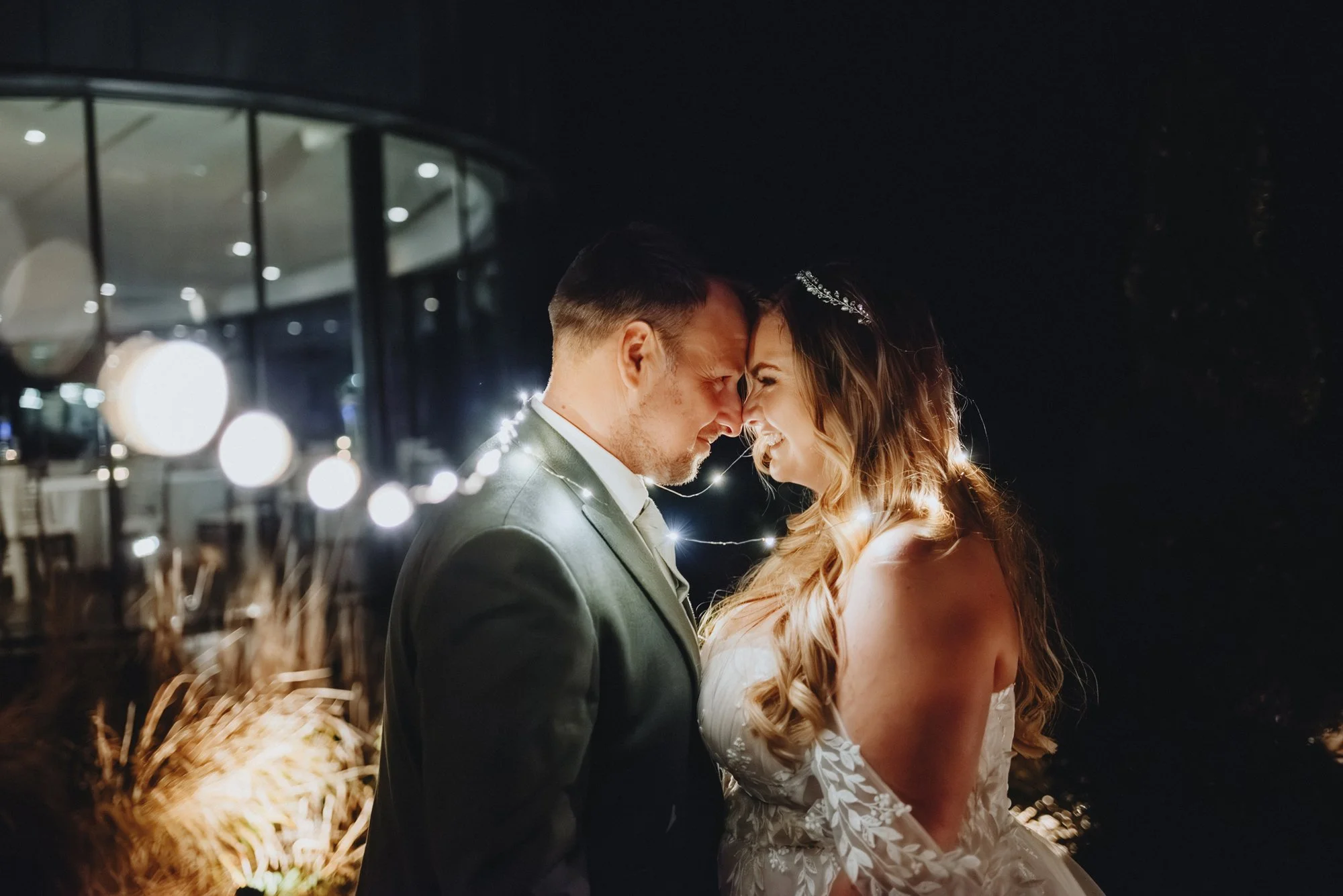 A couple on their wedding night with their foreheads touching, illuminated by string lights at night.