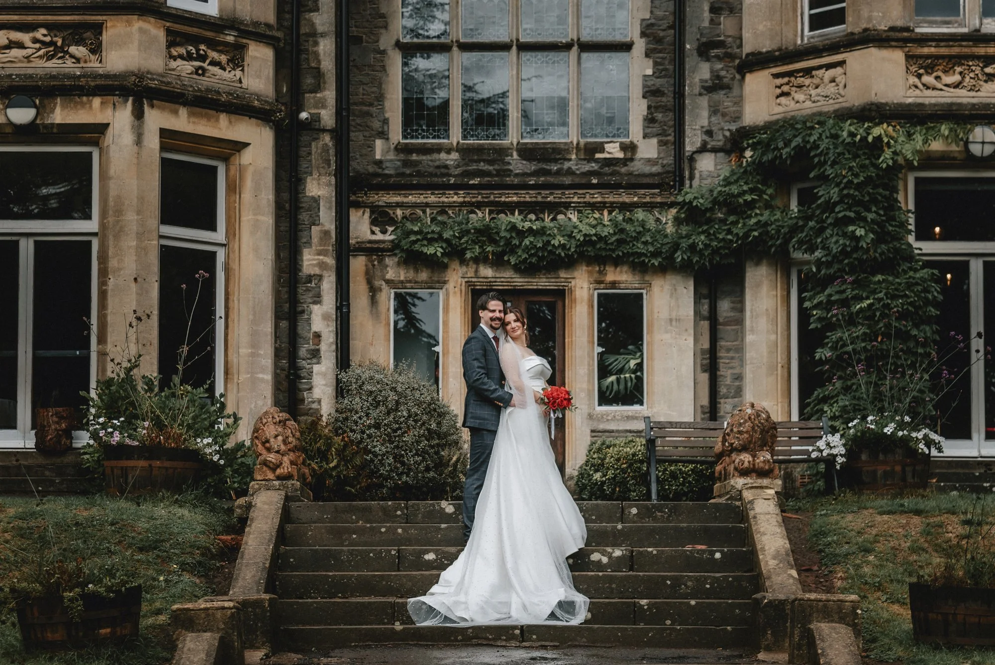 A bride and groom standing on steps outside a historic stone building with ivy and large windows, celebrating their wedding.