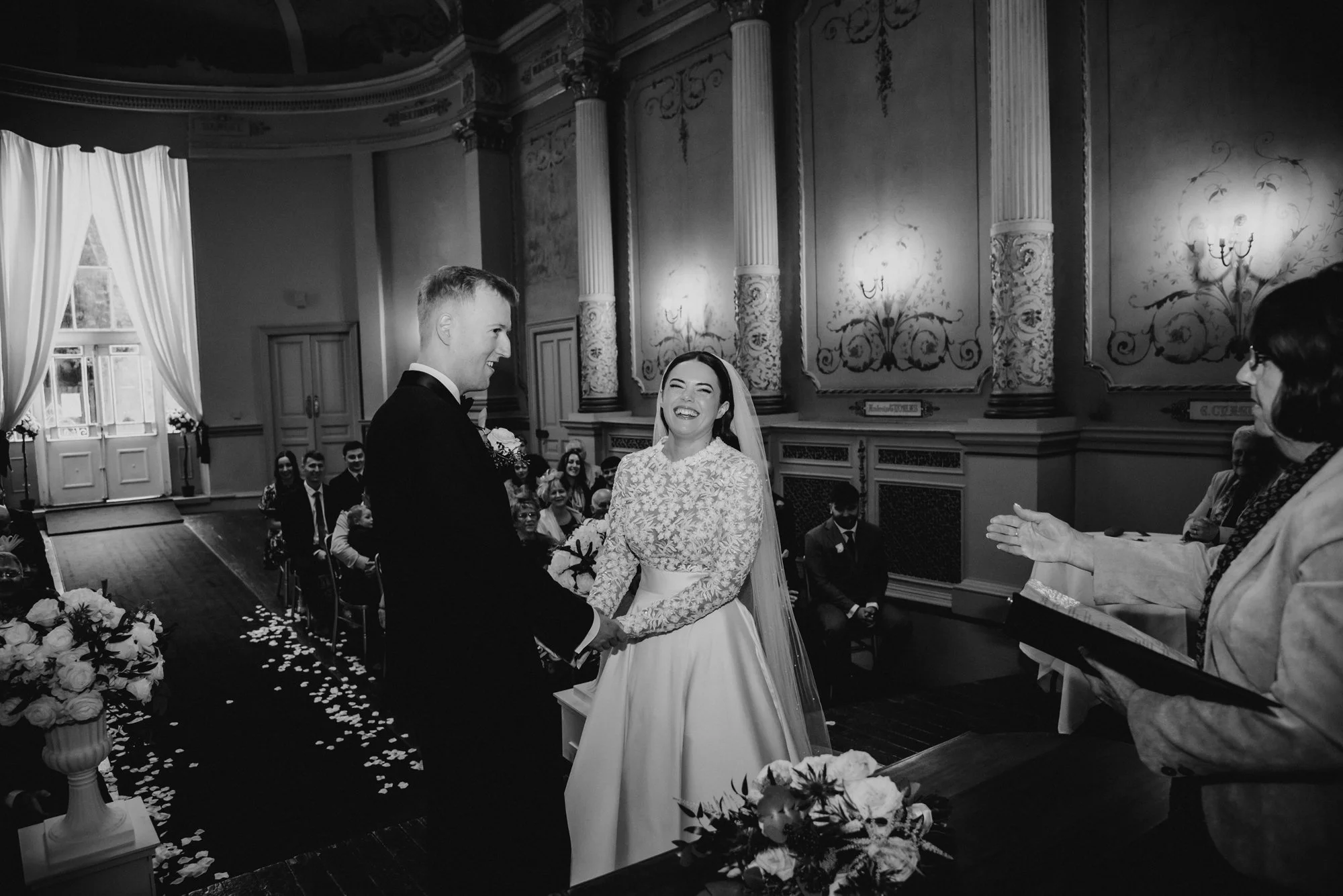 Black and white photo of a wedding ceremony with a bride and groom holding hands, smiling at each other, amid guests in an ornate, vintage interior. An officiant stands to the right.