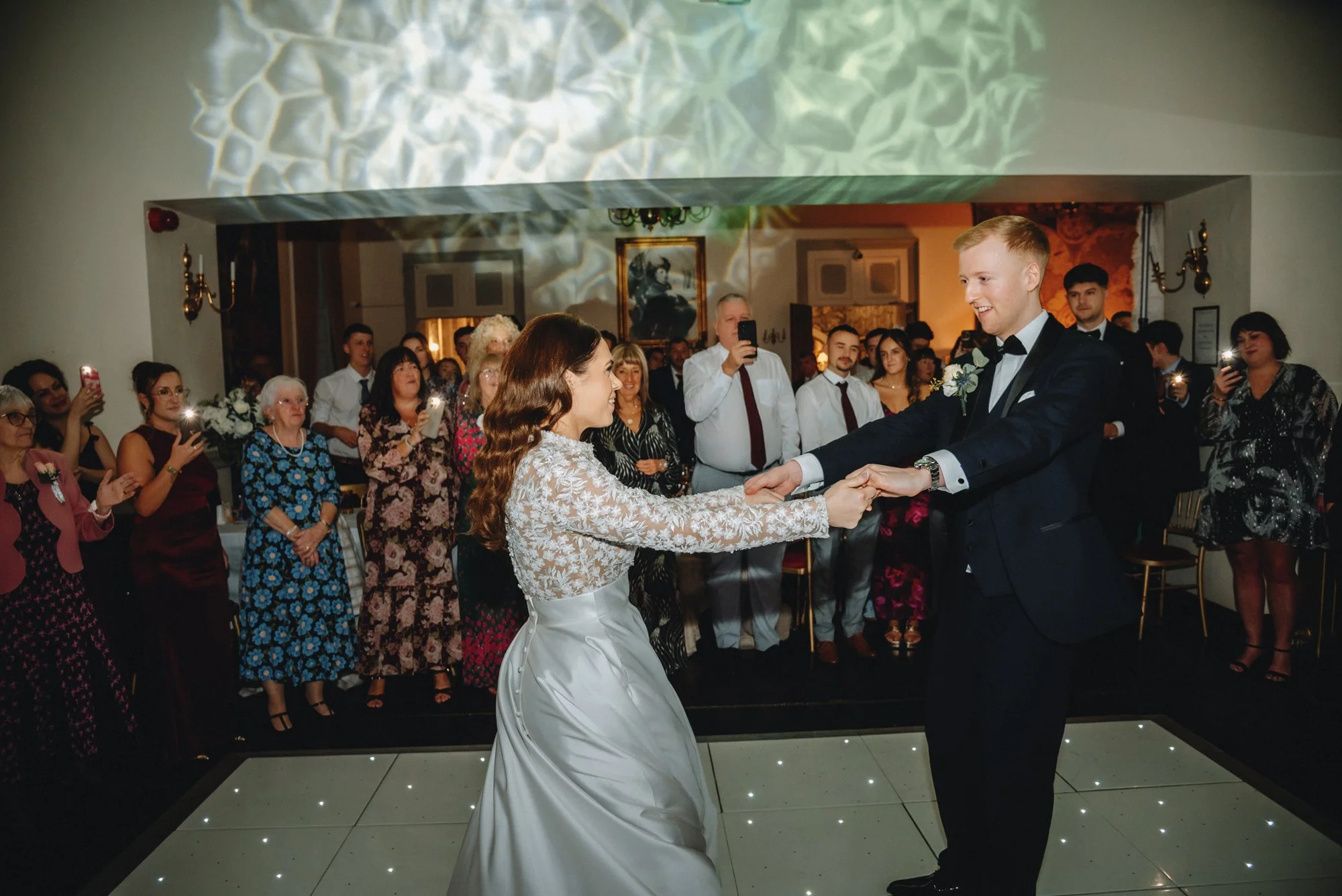 A bride and groom dancing at their wedding reception, with guests watching and taking photos in the background.