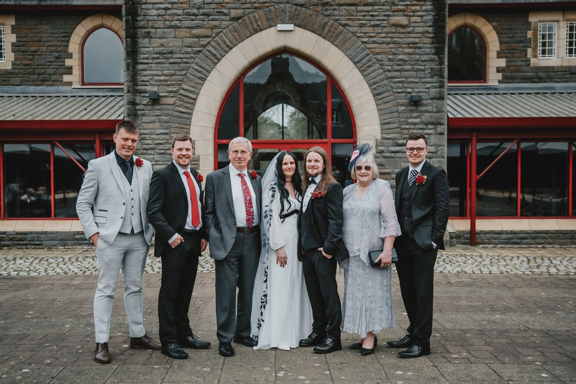 Group of seven people dressed in formal attire standing outside in front of a stone building with large arched windows during a wedding celebration.