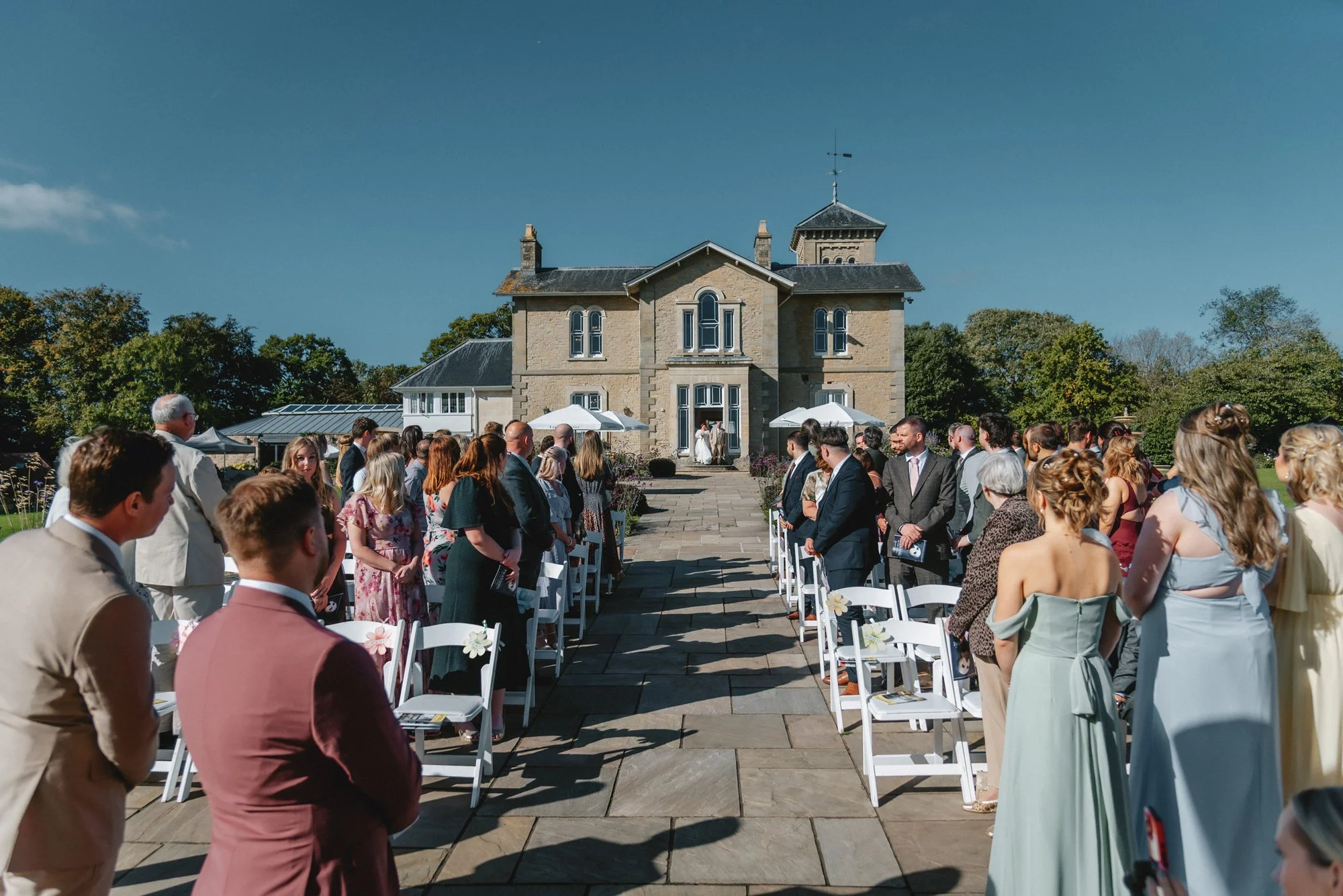 Outdoor wedding ceremony with guests standing on either side of the aisle, facing a bride and groom in front of a large stone house with a tower, under a clear blue sky.