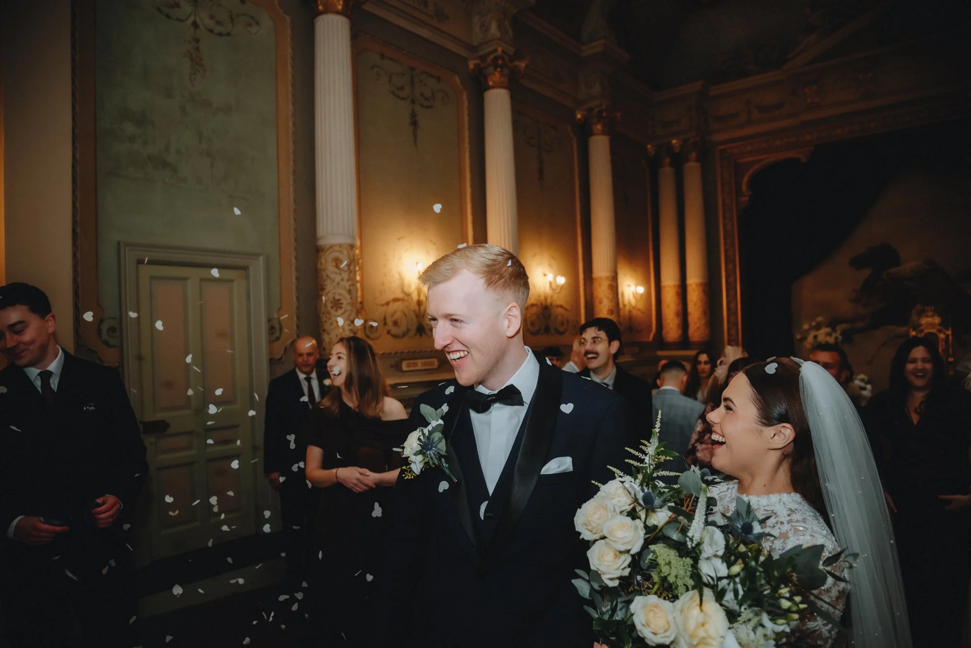 A newlywed couple at their wedding reception, the groom is smiling and wearing a tuxedo with a bow tie, and the bride is holding a bouquet of flowers, both surrounded by guests in an ornate, decorated hall, with confetti falling around them.