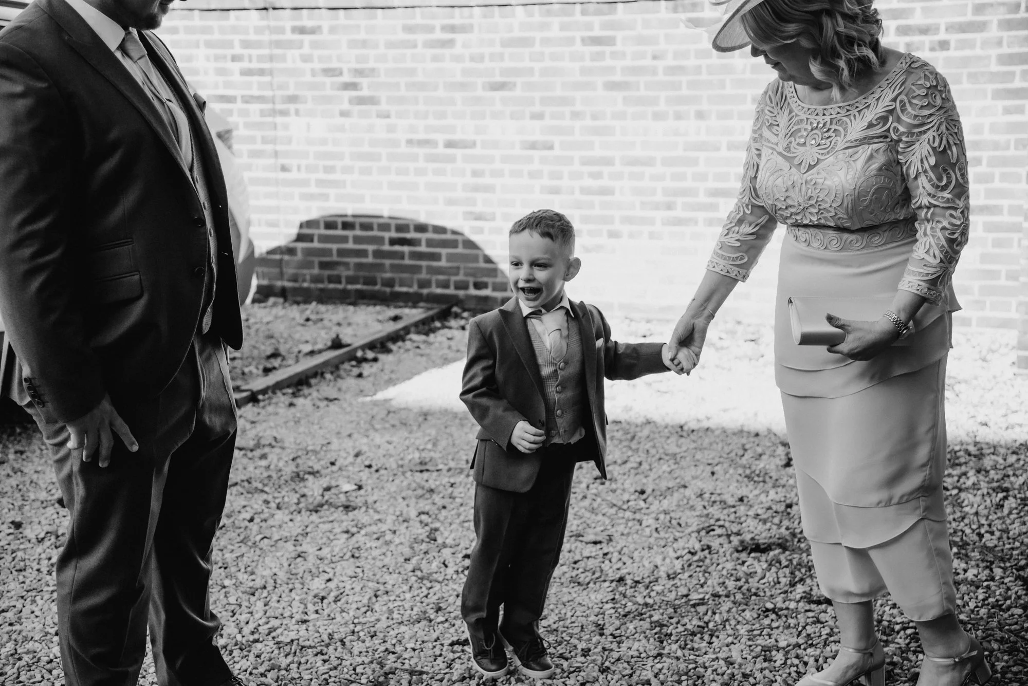 A young boy in a suit holding hands with a woman, smiling. Another man stands nearby. They are outdoors on gravel, with a brick wall in the background, dressed in formal attire.