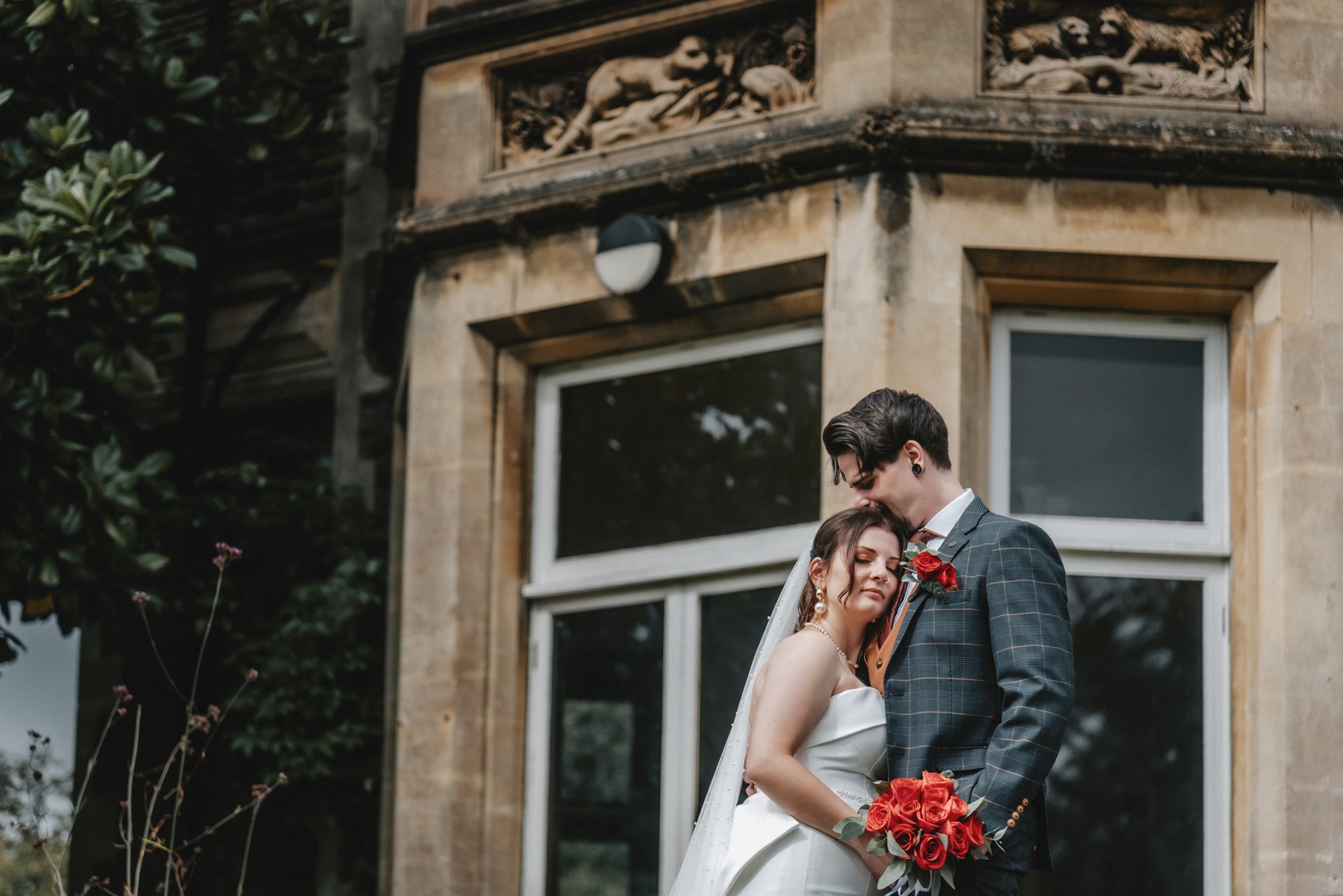A bride and groom embrace outdoors, standing close with eyes closed, in front of a stone building with decorative carvings and large windows. The bride holds a bouquet of red roses, and the groom wears a checkered suit with a red rose boutonniere.