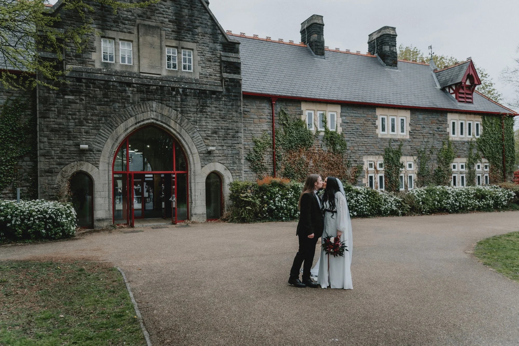 A couple in wedding attire sharing a kiss outside a historic stone building with a large arched glass doorway, surrounded by greenery and flowers.