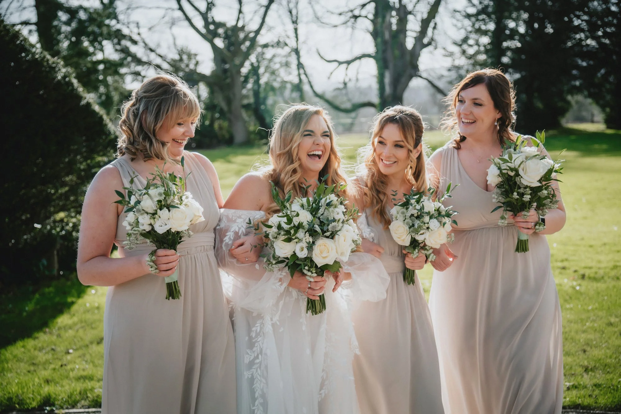 Bride and four bridesmaids laughing and holding bouquets during a wedding outdoors on a sunny day.