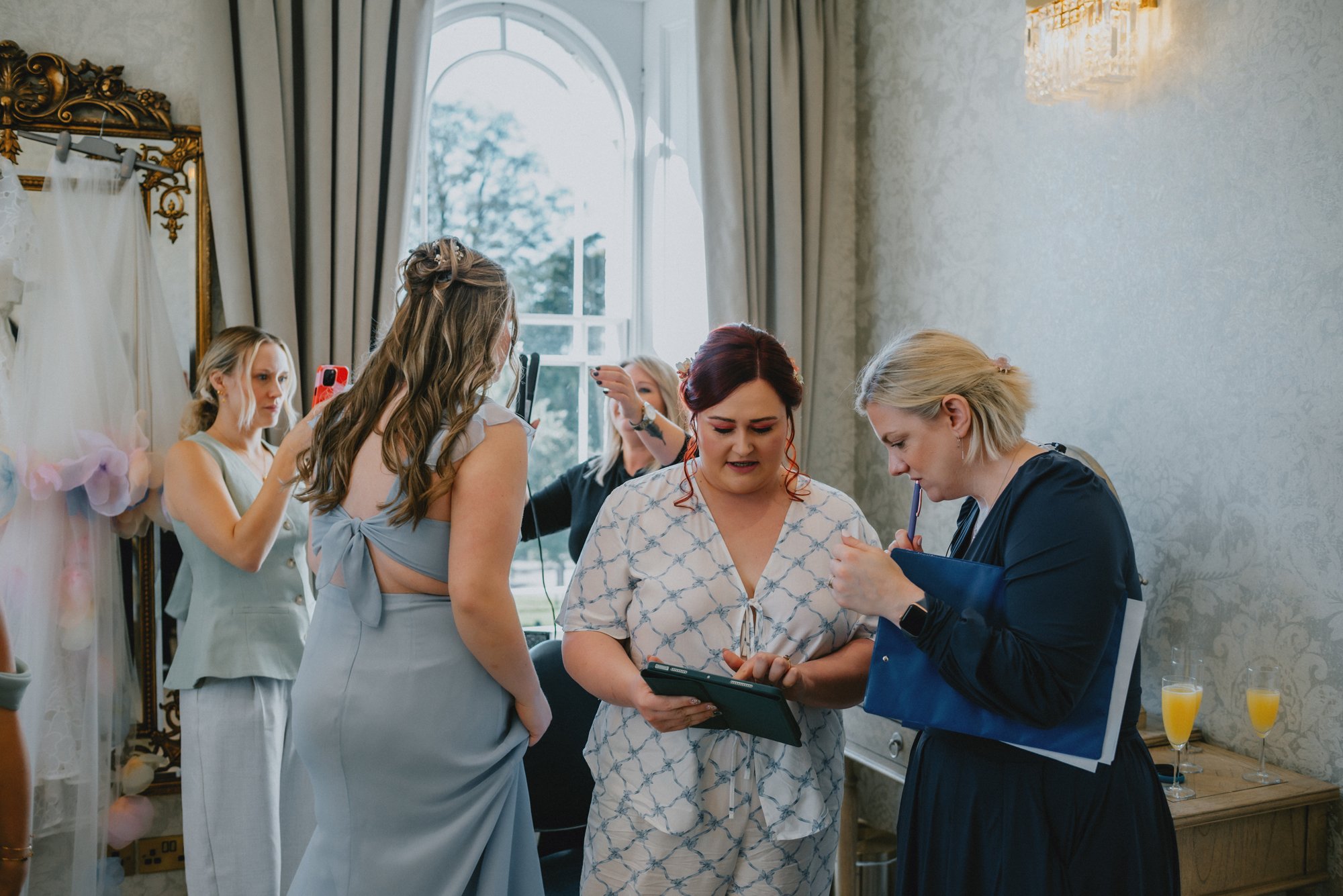 Group of women preparing for a wedding in a room with a large window, mirror, and drinks on a table.