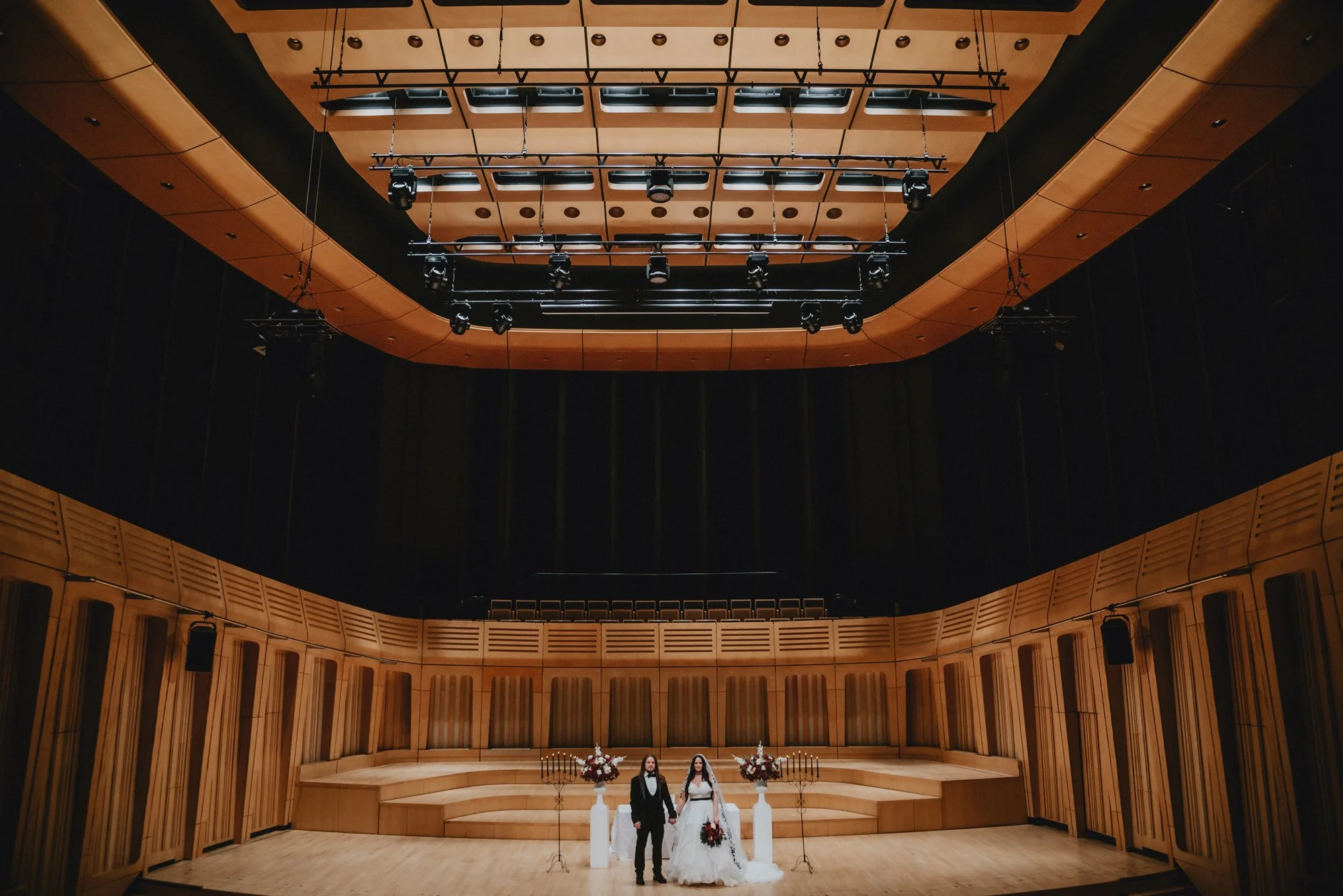 A bride and groom standing on a stage in a large, empty concert hall, decorated with floral arrangements and candelabras.