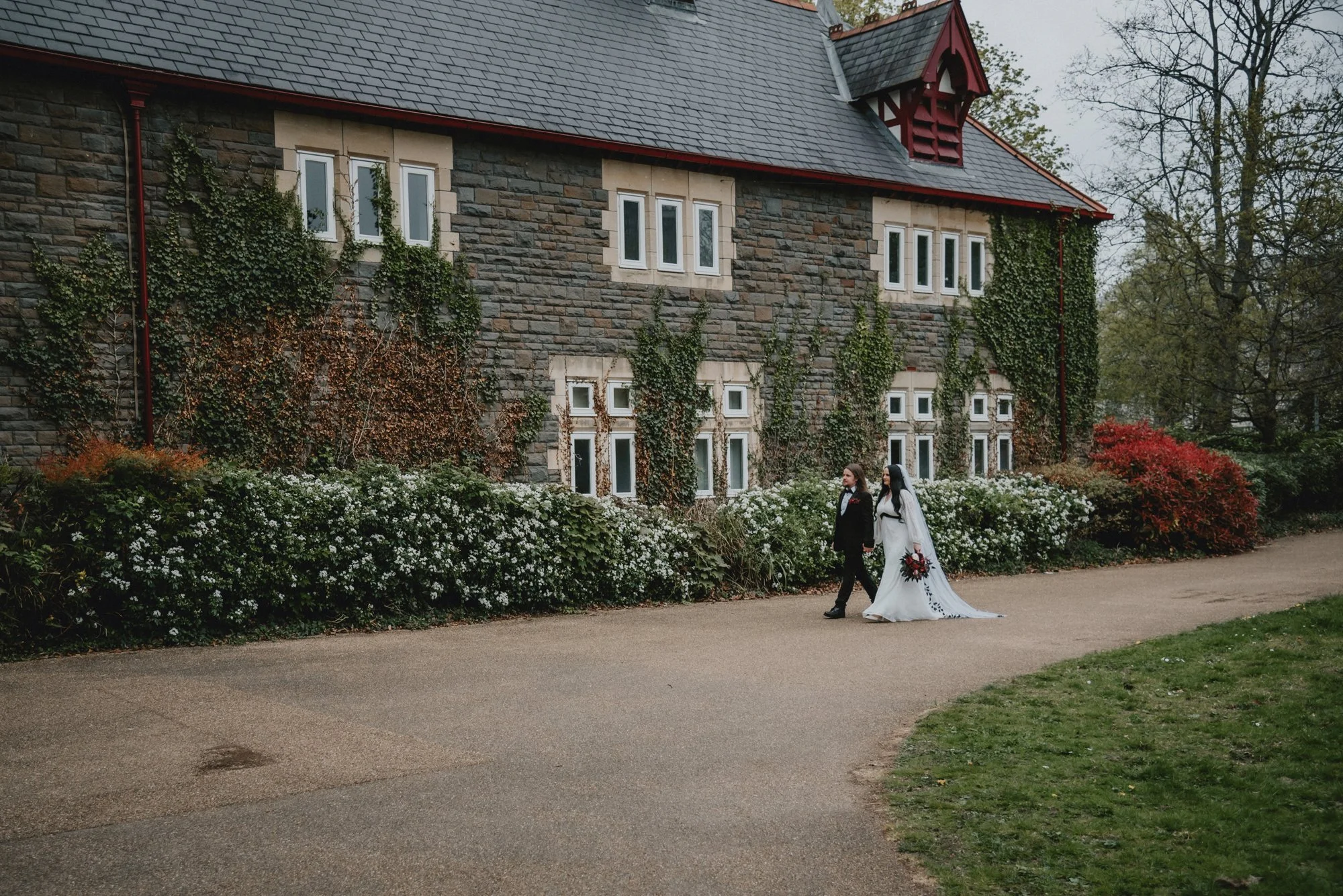 A bride and groom walking together outdoors in front of a stone building with ivy-covered walls and many windows. The bride is wearing a white wedding dress and holding a bouquet, while the groom is dressed in a black suit and tie.
