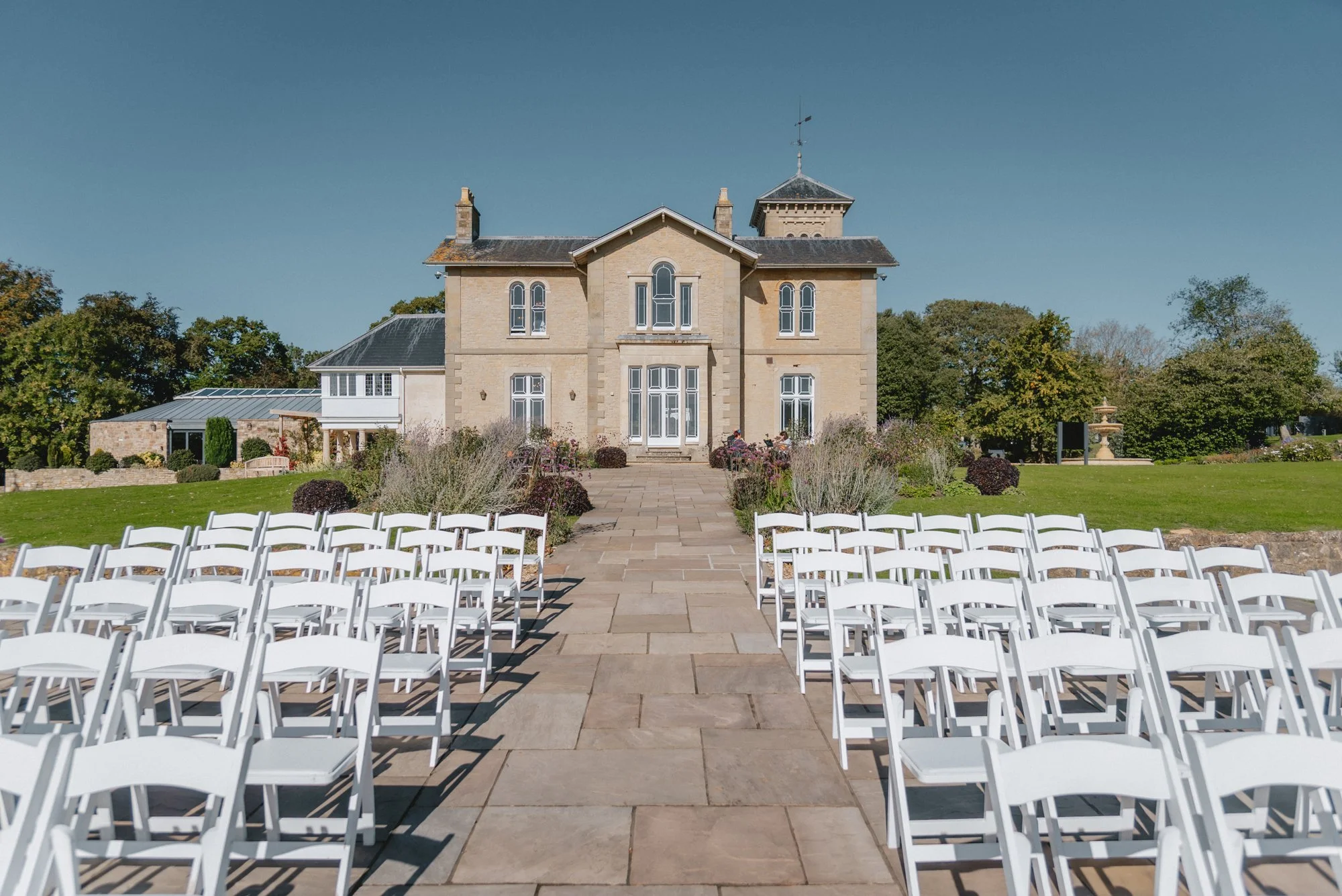 Outdoor wedding ceremony setup with rows of white chairs on a stone pathway leading to a large historic house, with a garden and trees in the background.