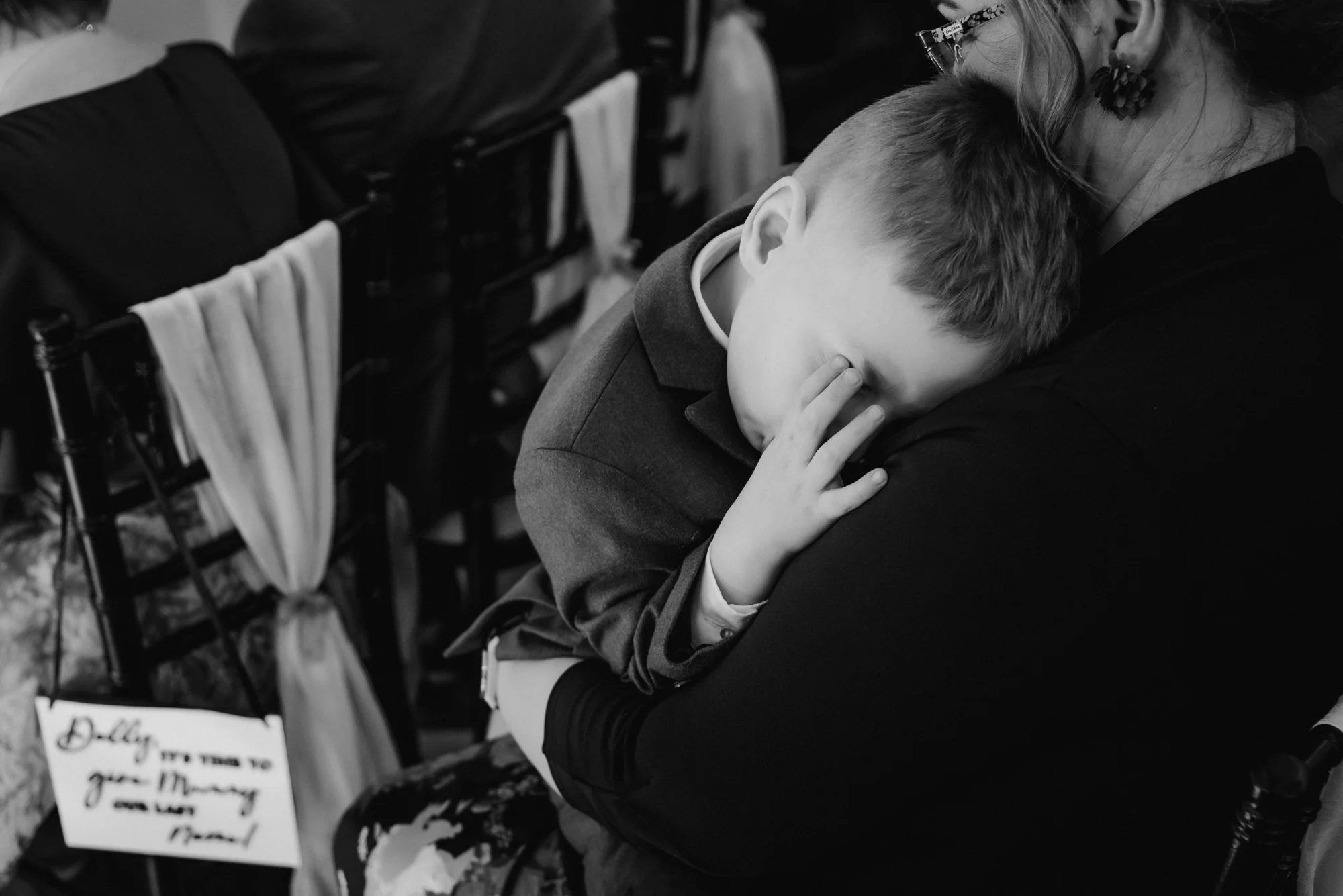 A young boy resting his head on an adult woman’s shoulder, appearing tired or sad, in a setting with chairs and a sign that says "Dolly, you'll free the money and let me..."