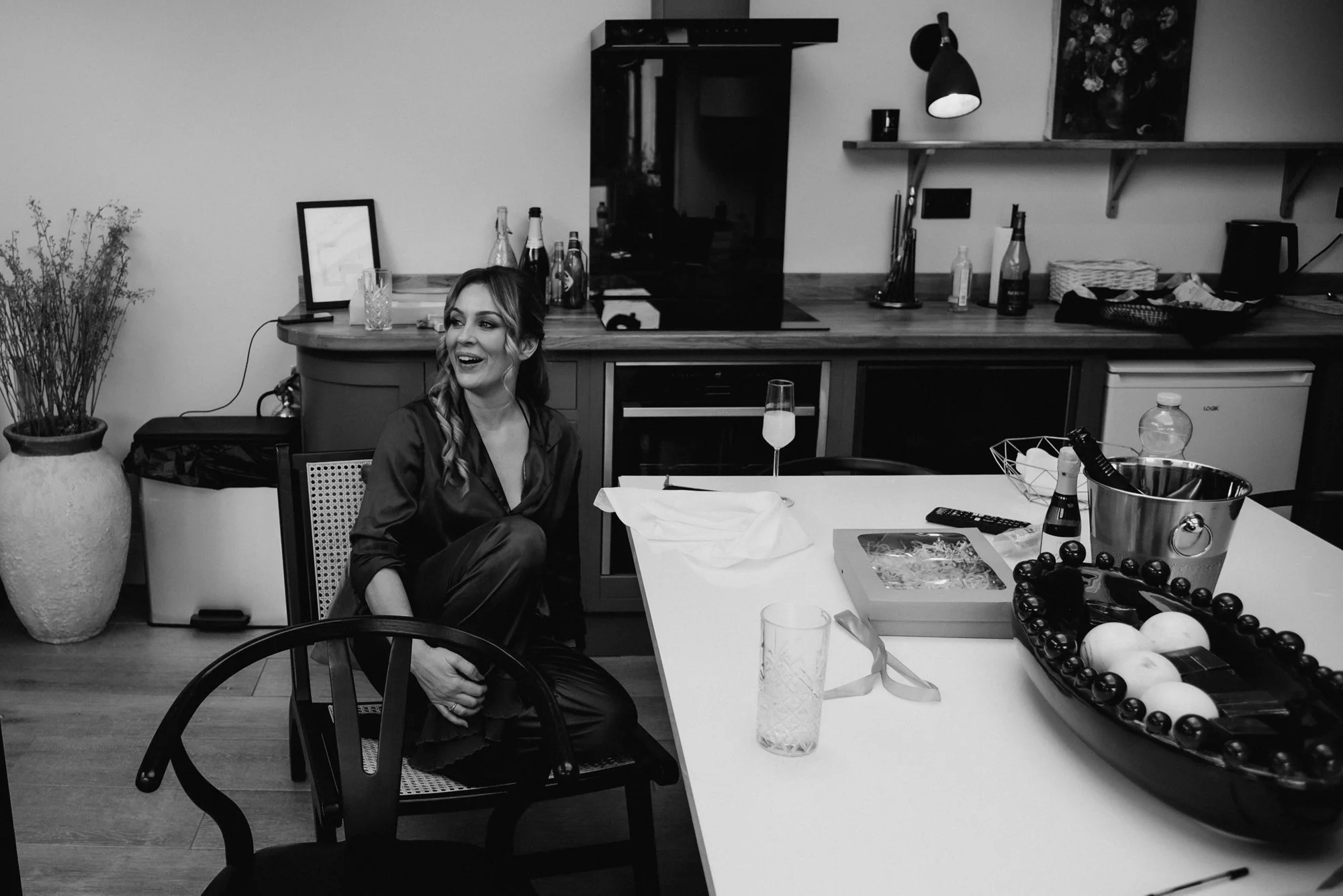 A woman sitting happily on a chair in a kitchen, smiling and looking to her left. The kitchen has a counter with various bottles, a glass with a drink, and a plate. On the table in the foreground, there are glasses, a wrapped gift, a bottle of champa