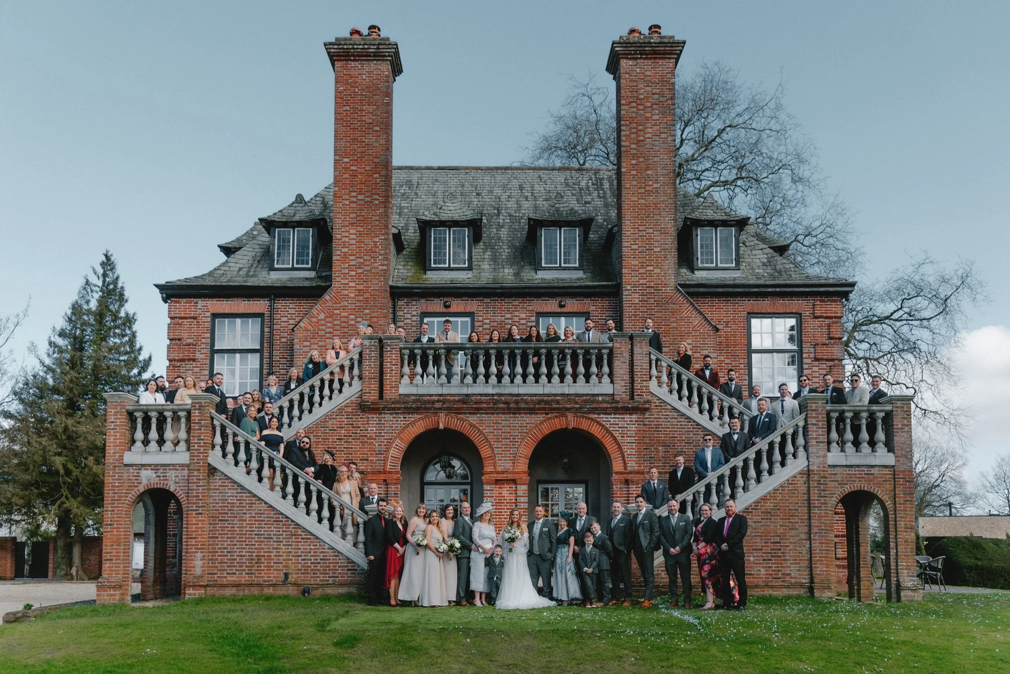 A large group of people dressed in formal and wedding attire standing in front of a historic brick mansion with an elaborate staircase and outdoor terrace.