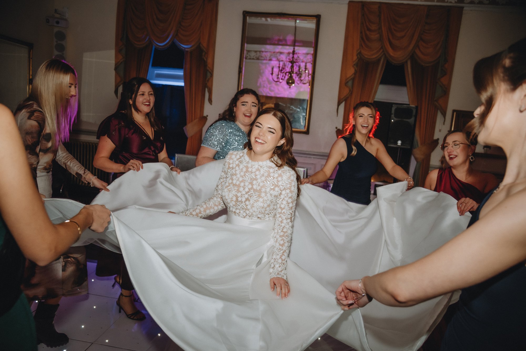 Women in formal attire holding the train of a bride as she sits and smiles during a wedding celebration.