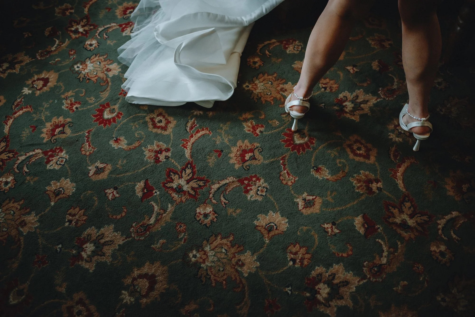 Close-up of a woman wearing white high heels with pearl ankle straps, standing on a patterned green, red, and beige rug. Part of a white dress is visible, resting on the floor.