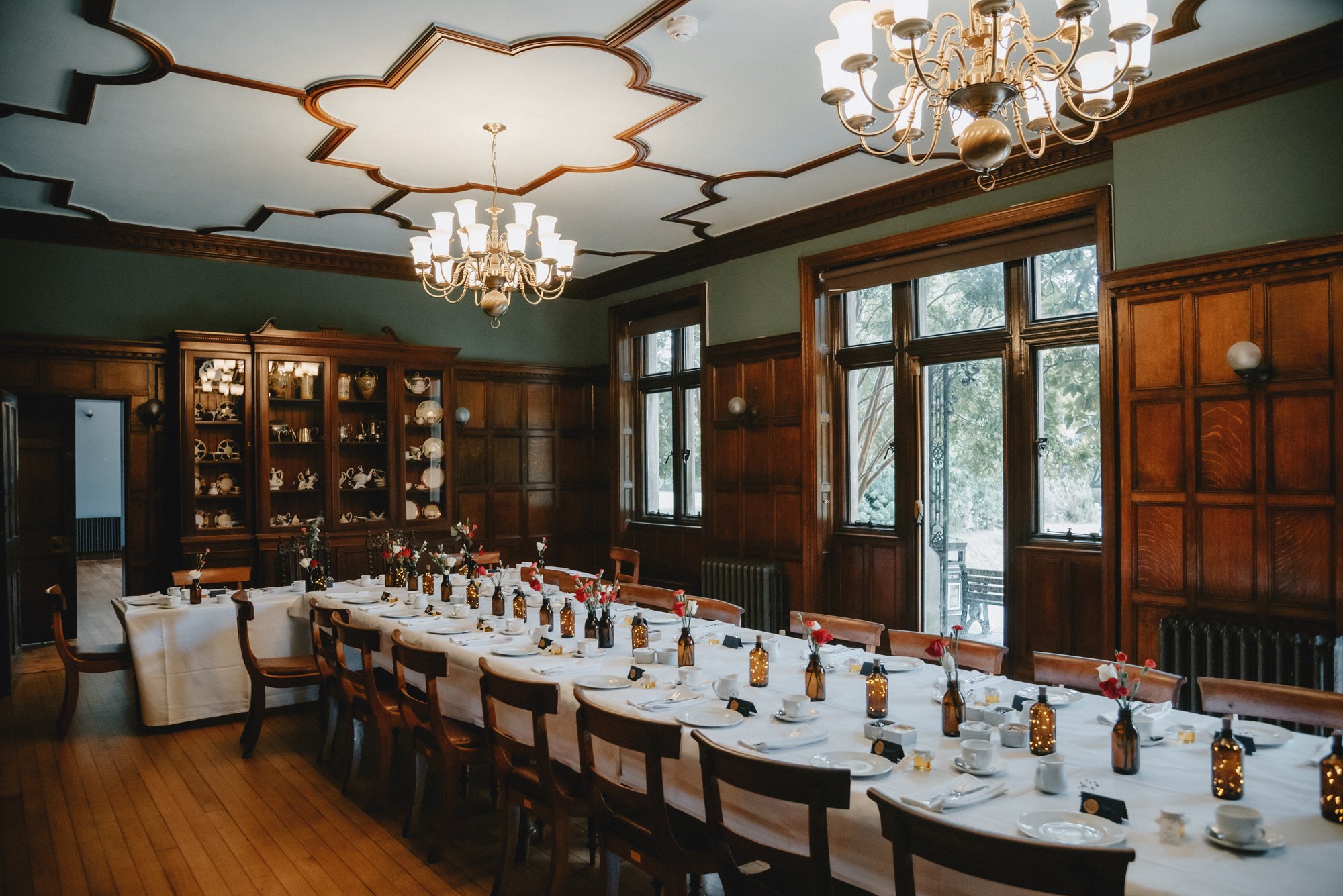 A formal dining room with a long table set for a meal. The table has a white tablecloth, plates, cups, and small bottles with flowers. There are wooden chairs around the table, and large windows with wooden frames letting in natural light. The room h