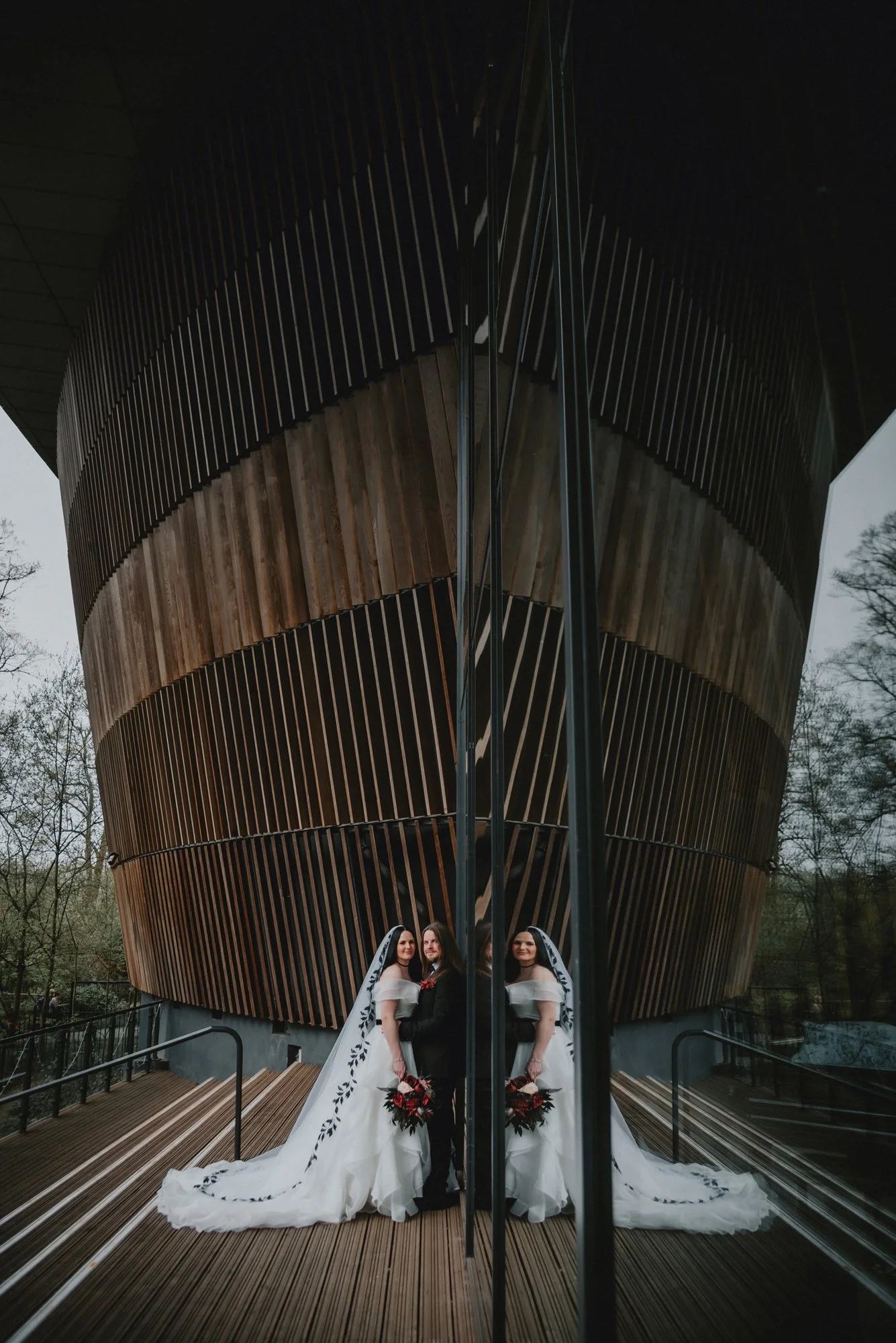 Wedding couple standing in front of a modern wooden building, captured through a mirror creating a symmetrical reflection.