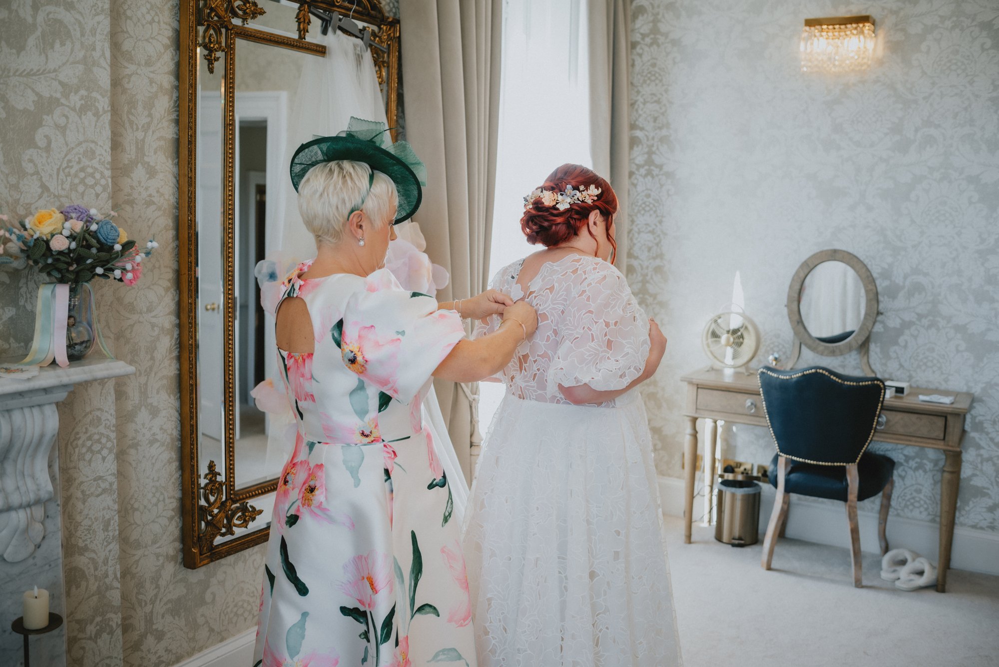 An elderly woman helping a bride into her wedding dress in a bright, elegantly decorated room with floral wallpaper, a mirror, and a vanity table.