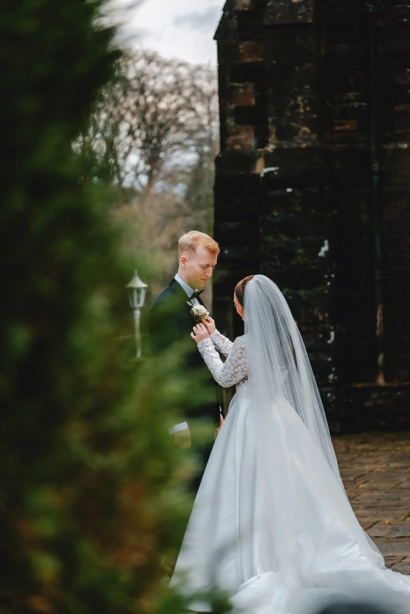 A bride and groom sharing a moment outside during their wedding, with the bride in a white gown and veil, and the groom in a tuxedo, near an old brick building and surrounded by greenery.