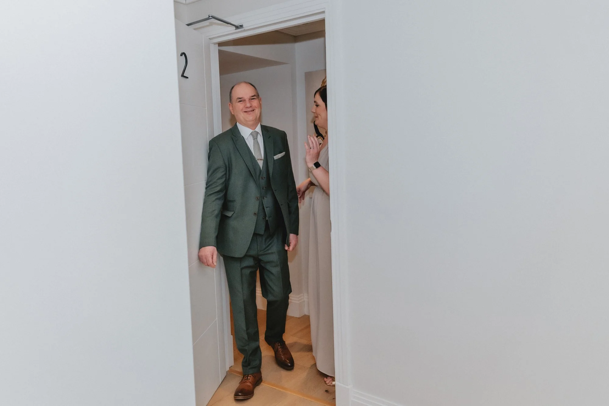 A man in a green suit and a woman in a beige dress standing in a doorway and smiling at each other.