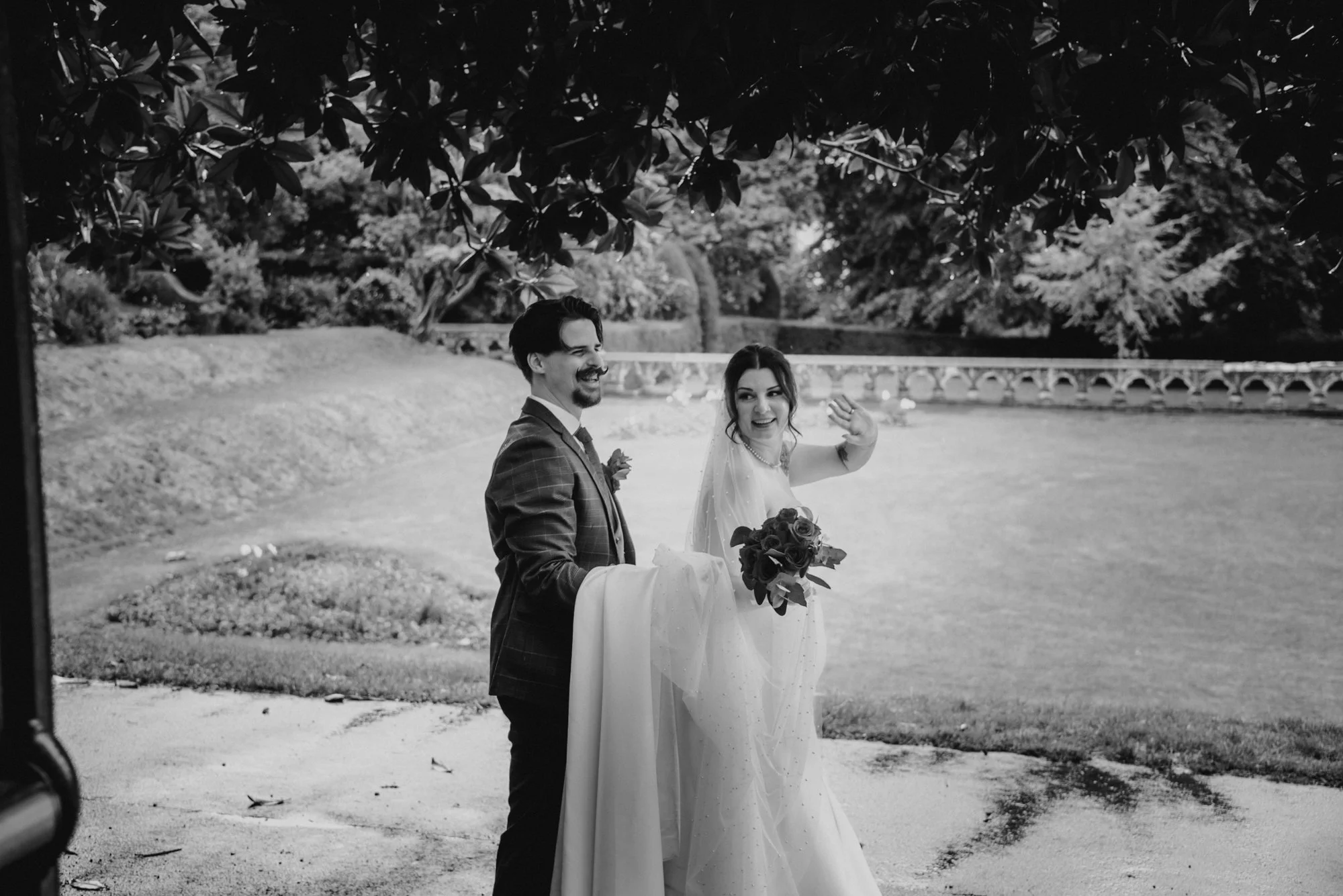 Black and white photograph of a bride and groom outdoors, with the bride waving and holding a bouquet, under trees with a stone bridge in the background.
