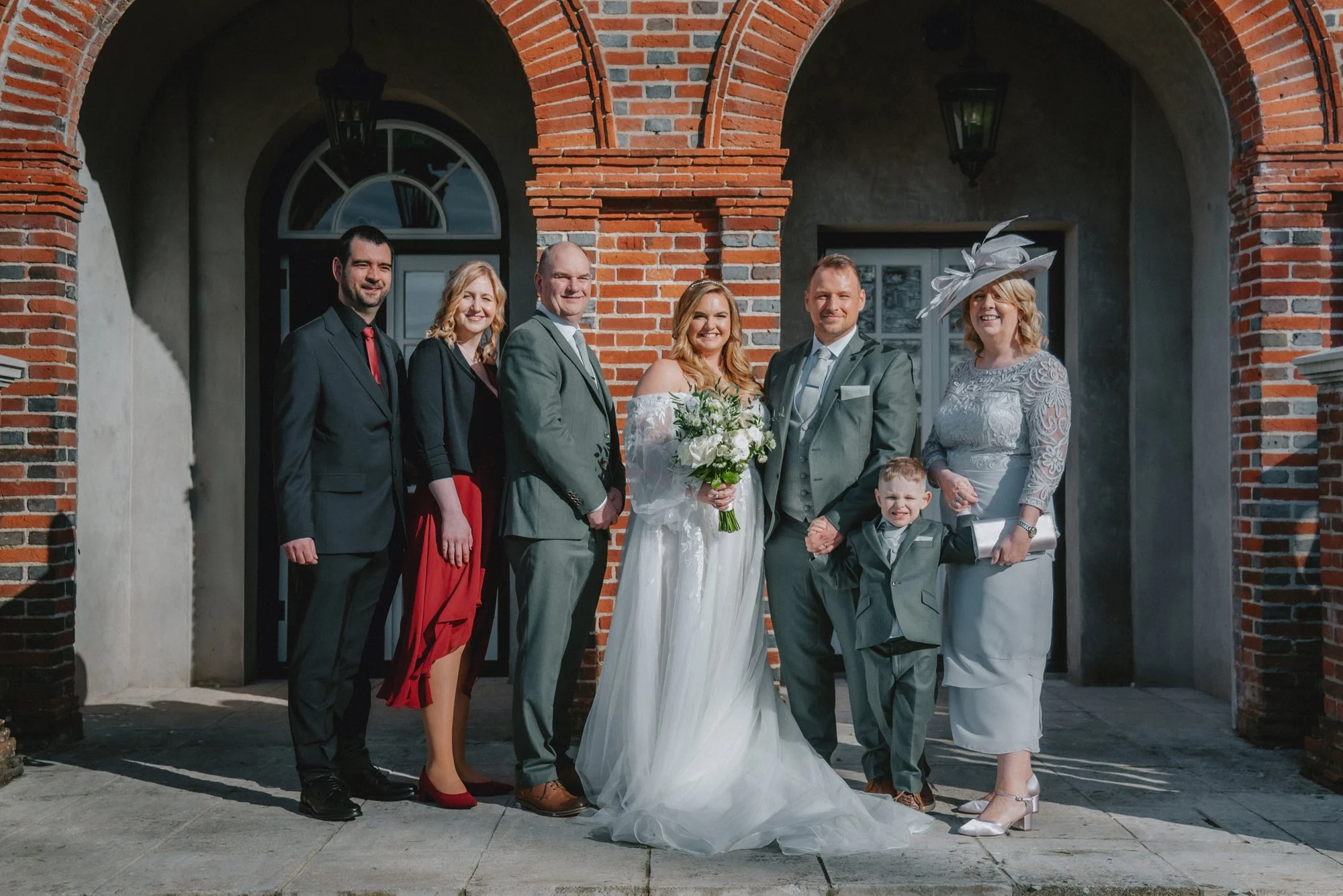 A group photo of a wedding party outside a brick building with arched entryways, featuring seven people dressed in formal attire, including the bride in a white gown holding a bouquet, and the groom in a gray suit holding hands with a young boy.