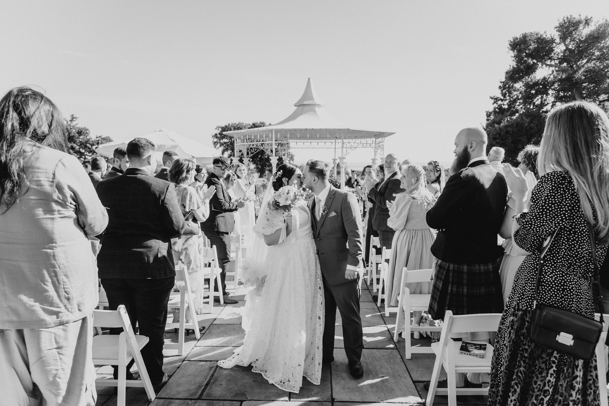 A black-and-white photo of a wedding ceremony outdoors with the bride and groom kissing, surrounded by seated and standing guests, under a decorative gazebo.