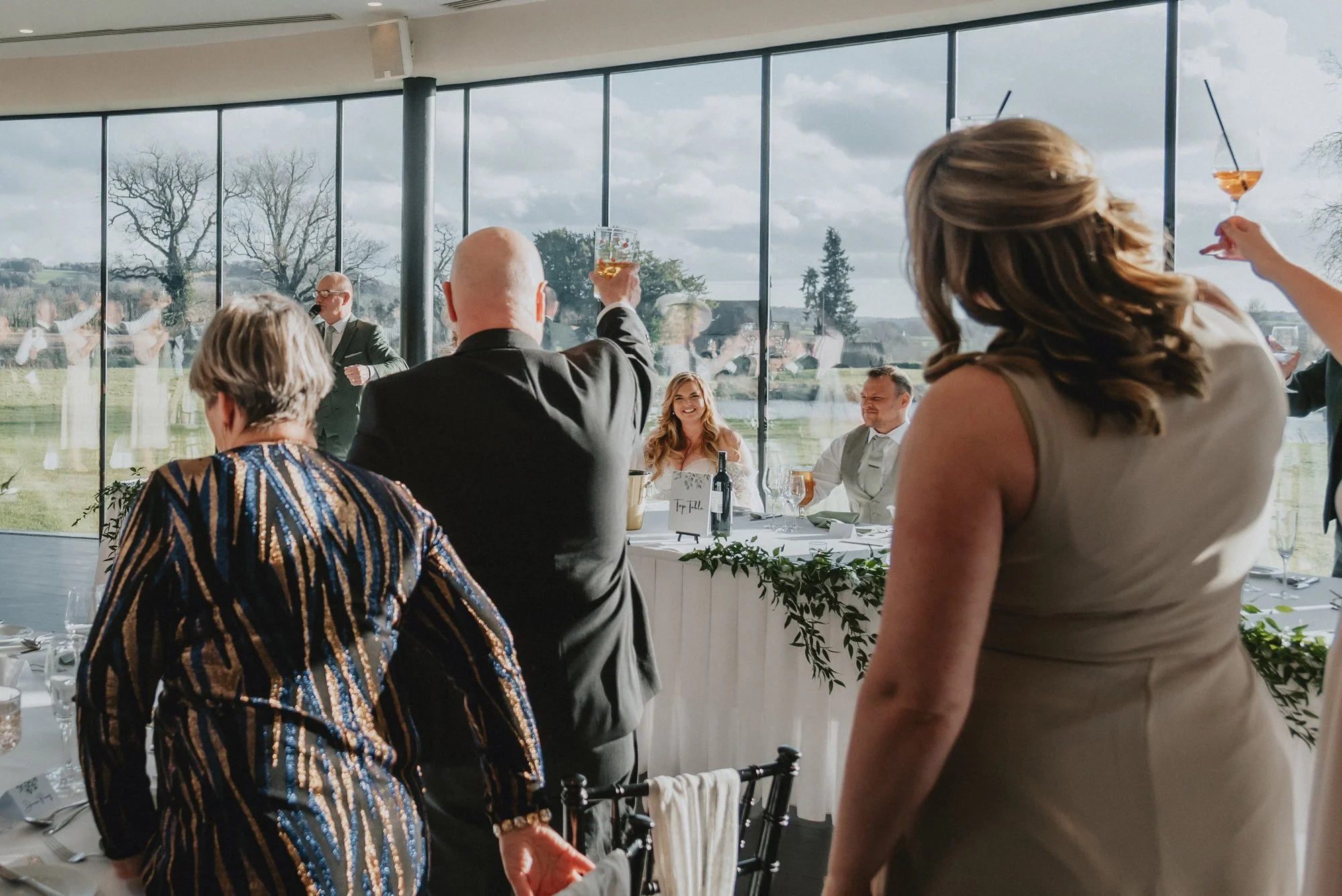 People raising glasses in toast at a wedding reception with large window showing outdoor scenery