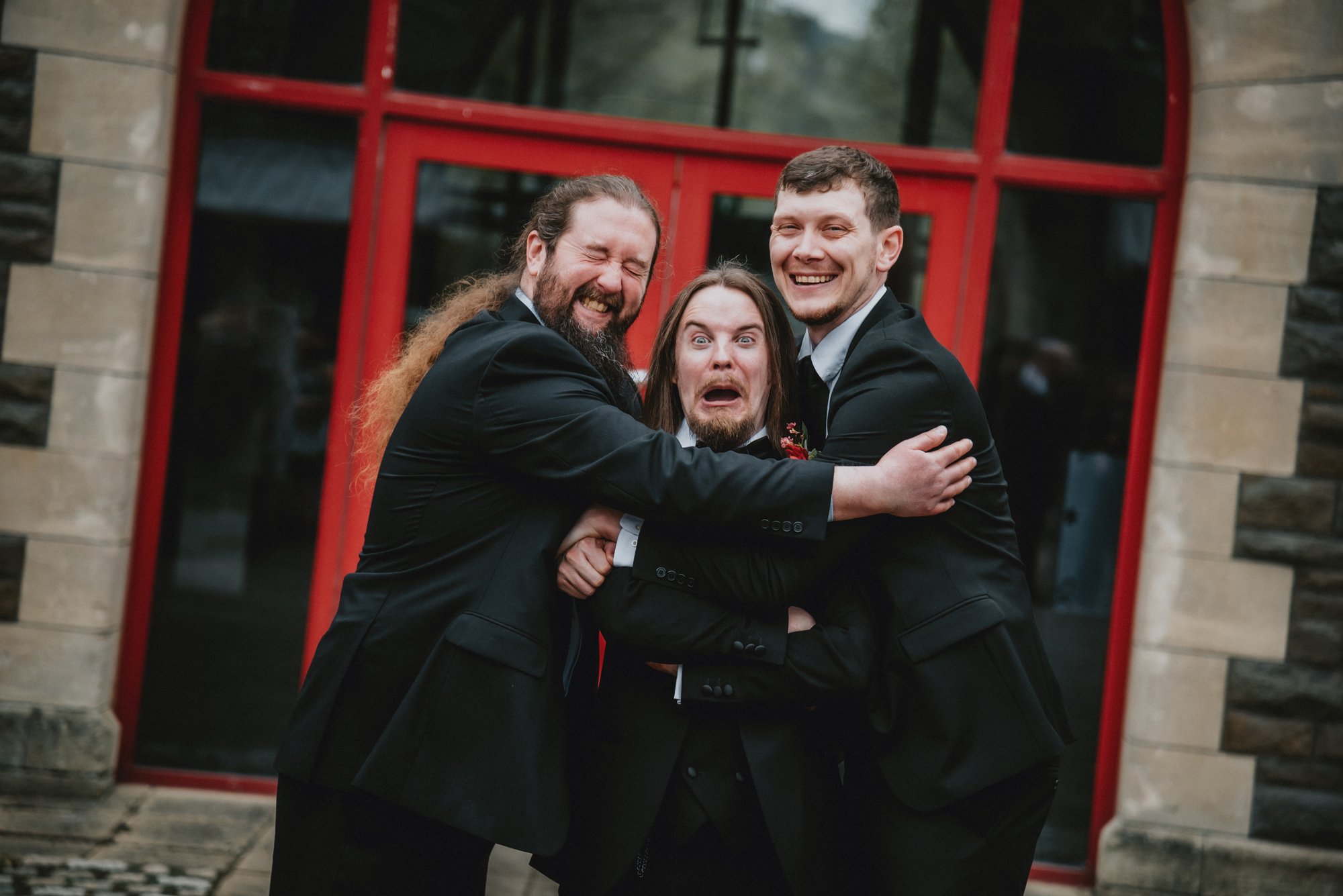 Three men in tuxedos hugging and making funny faces in front of a building with red-framed glass doors.