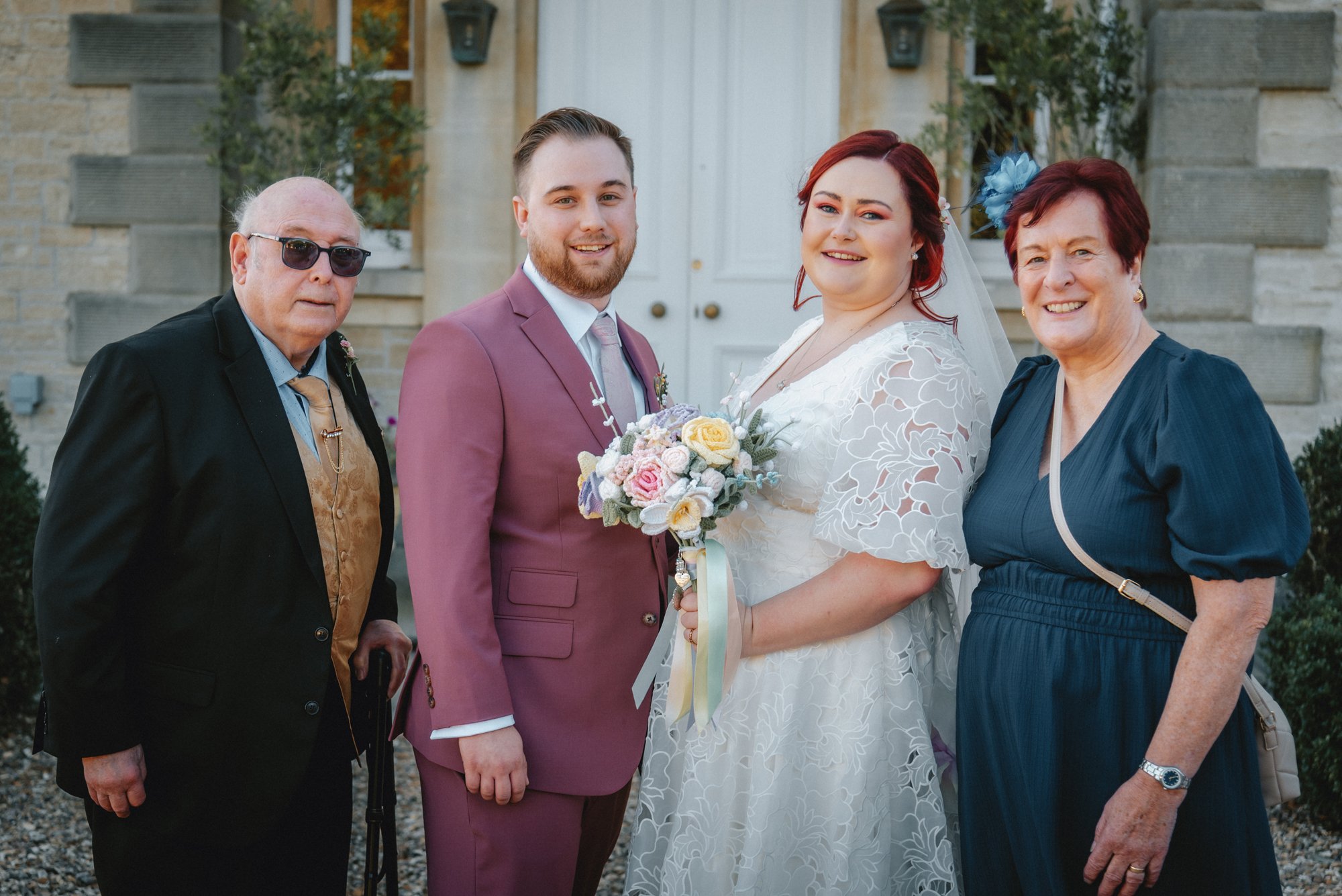 A group of four people standing outdoors in front of a stone building, including a bride and groom, with two older women, all smiling at the camera.