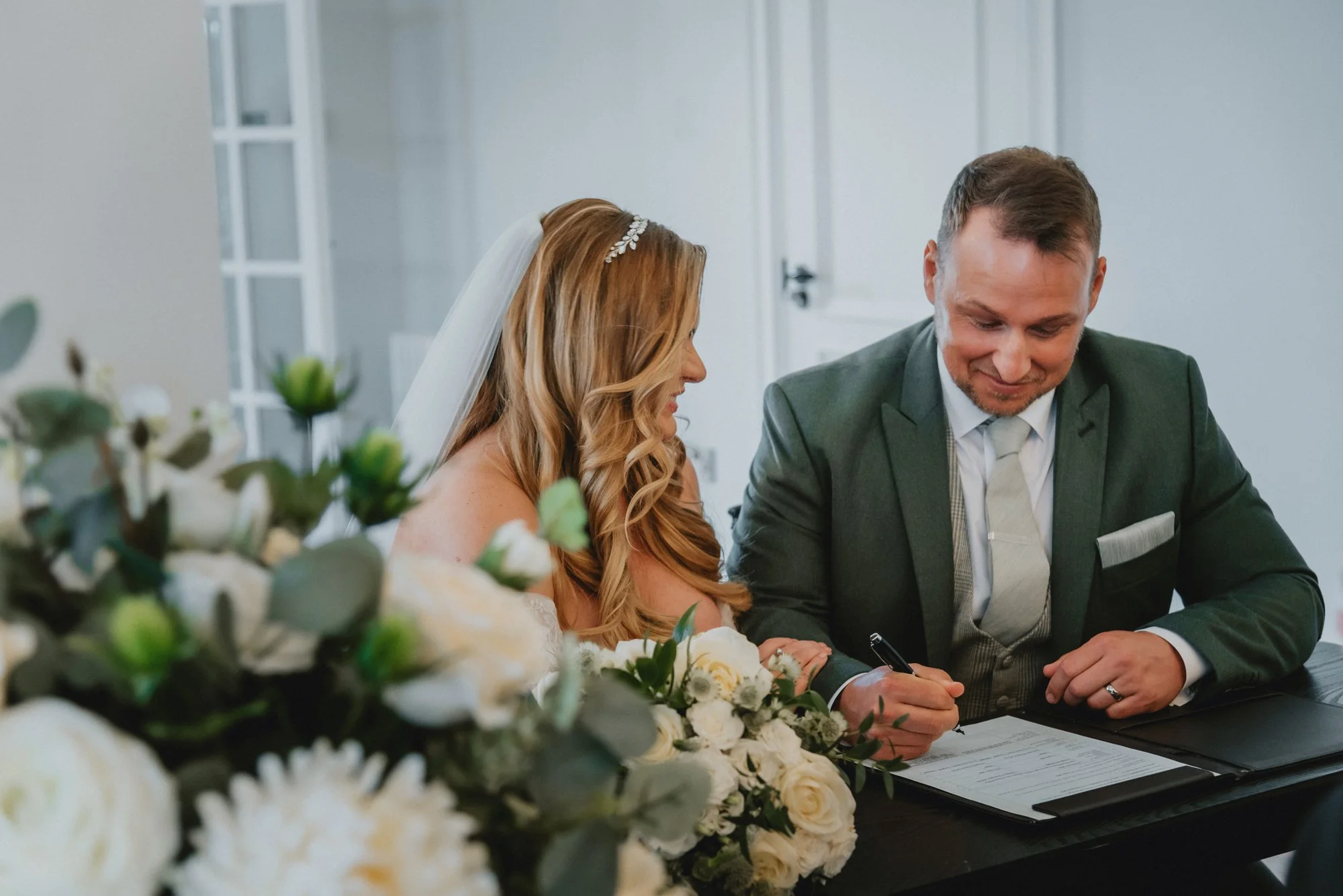 A bride with long red hair, wearing a white veil and dress, sits next to a groom with short brown hair, in a grey suit, signing a marriage license at a table decorated with white flowers.