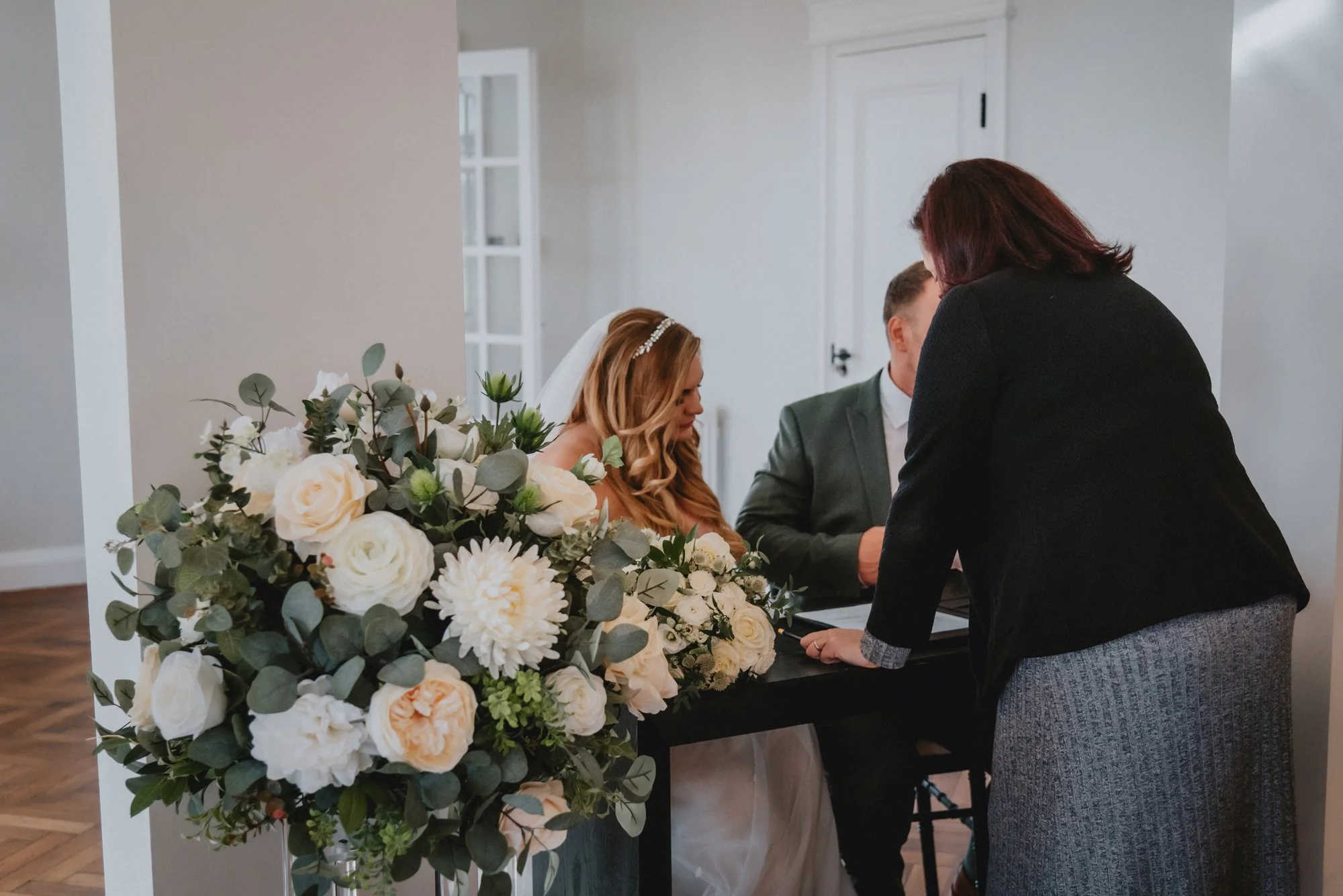 A bride and groom signing a document during their wedding ceremony, with a woman assisting them, in a room decorated with a large floral arrangement of white and cream flowers and greenery.