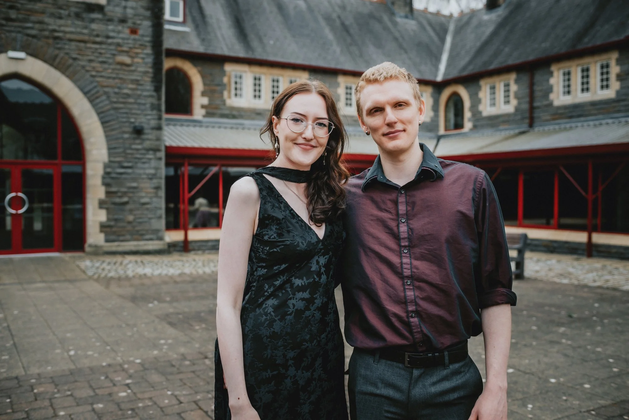 A young woman and man standing close together outdoors in front of a stone building with arched windows and red accents.