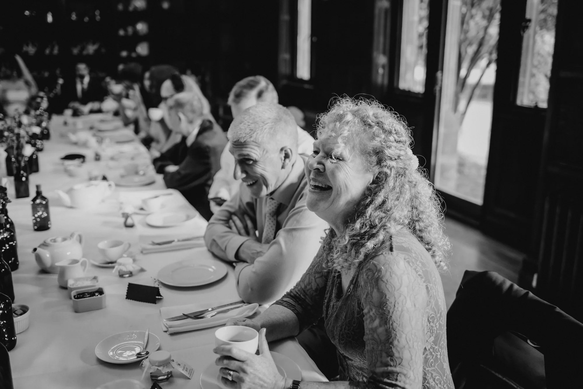 A group of older adults sitting at a long dining table, smiling and enjoying a meal together, in a room with large windows passing natural light.