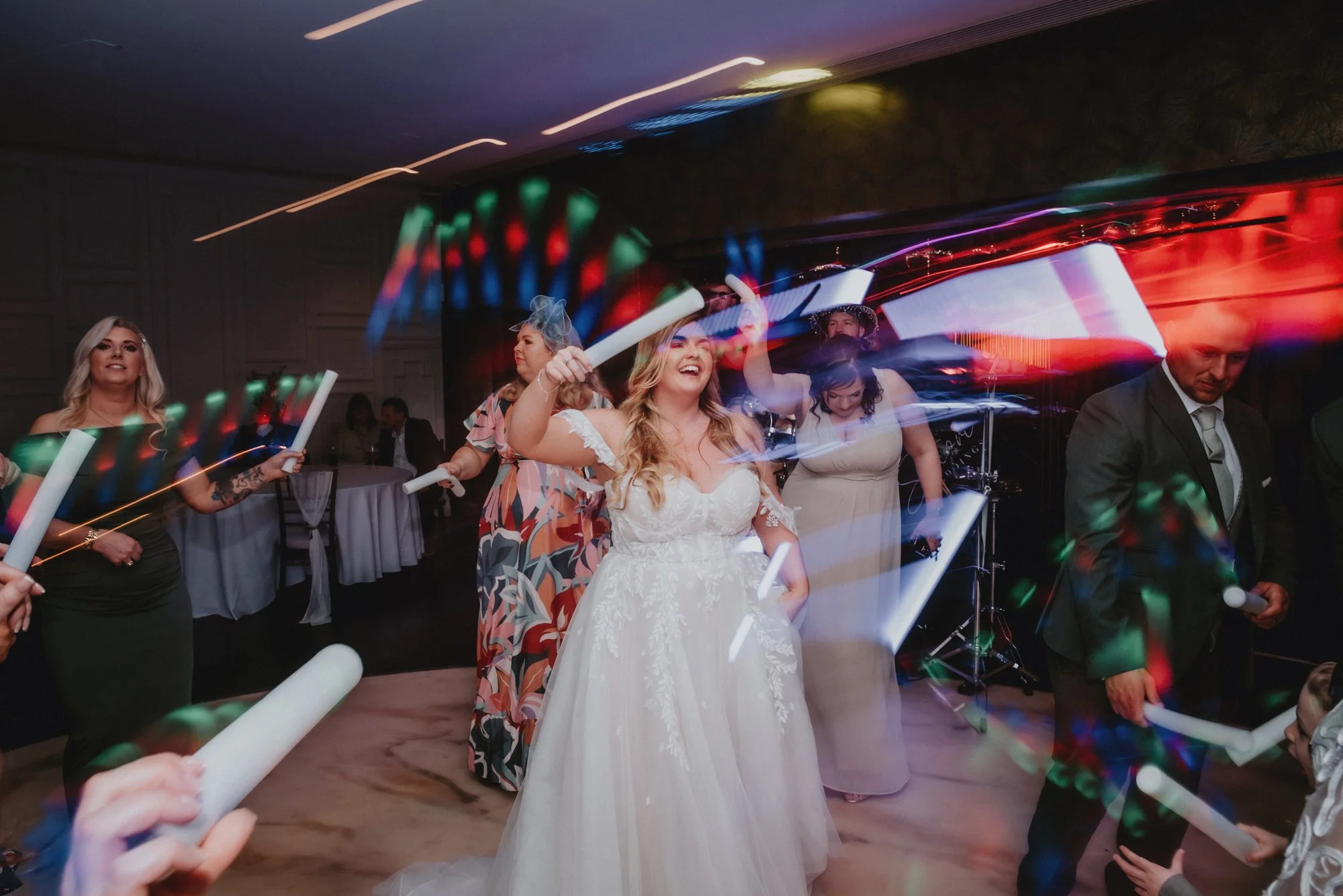 Wedding reception with guests dancing, smiling, and holding glow sticks in a dimly lit room with colorful light effects.