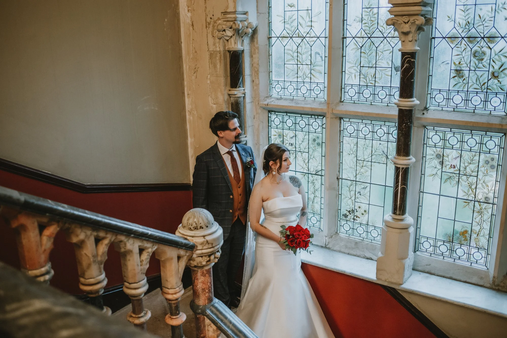 Bride and groom standing on a staircase near a large stained glass window, with the bride holding a bouquet of red roses, inside a historic building.