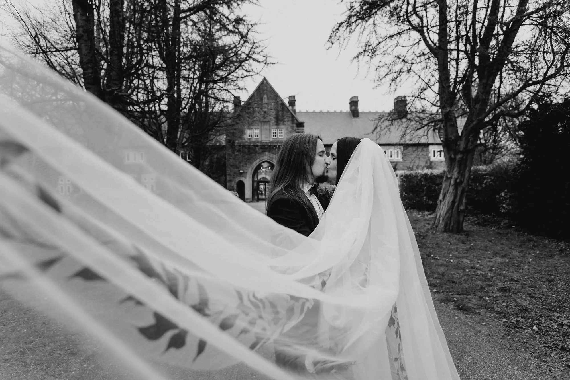 A black and white photo of a couple kissing, with the bride's veil flowing in the wind, outdoors in front of a large, historic building surrounded by trees.