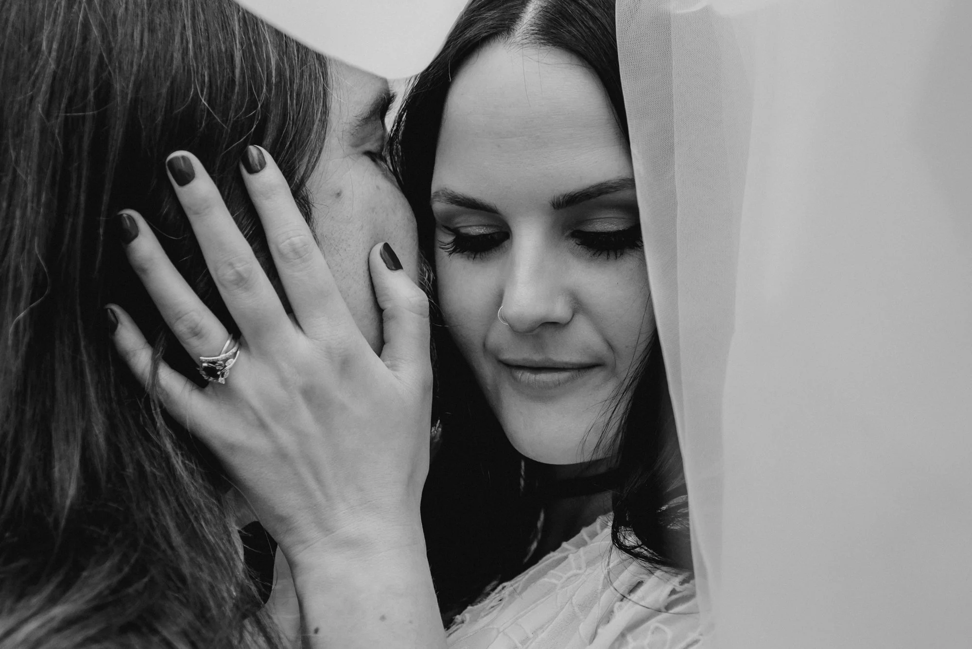 A black-and-white photograph of two women in close intimate pose, with one woman kissing the other on the cheek while the other woman softly smiles, wearing a veil.