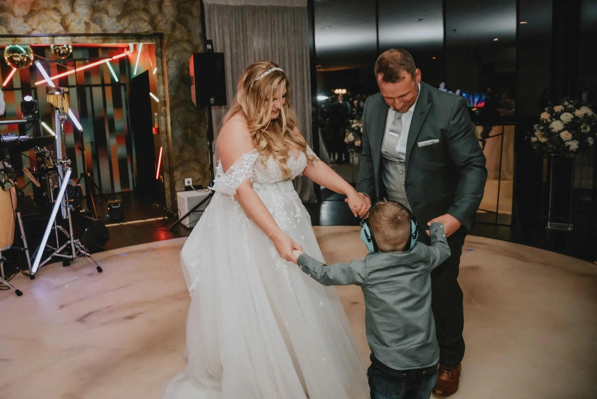 A bride and groom dancing with a young boy at a wedding reception, holding hands and standing on a dance floor.