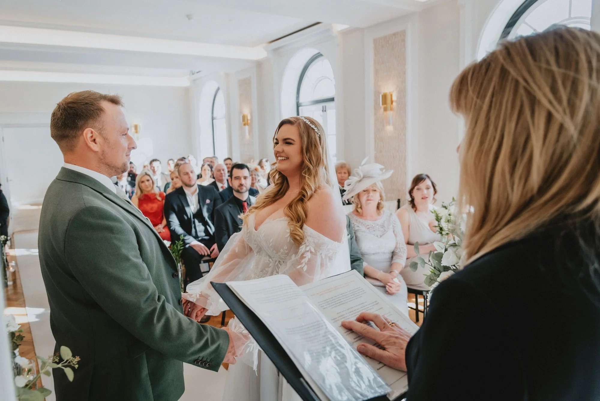 A bride and groom holding hands during their wedding ceremony in a bright, elegant room, with a female officiant reading from a book, surrounded by seated guests.