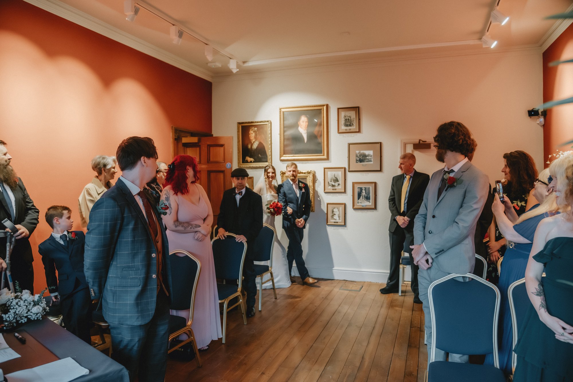 People gathered at a wedding ceremony in a room with framed artwork on the walls. The bride and groom stand at the front, surrounded by friends and family, some taking photos.