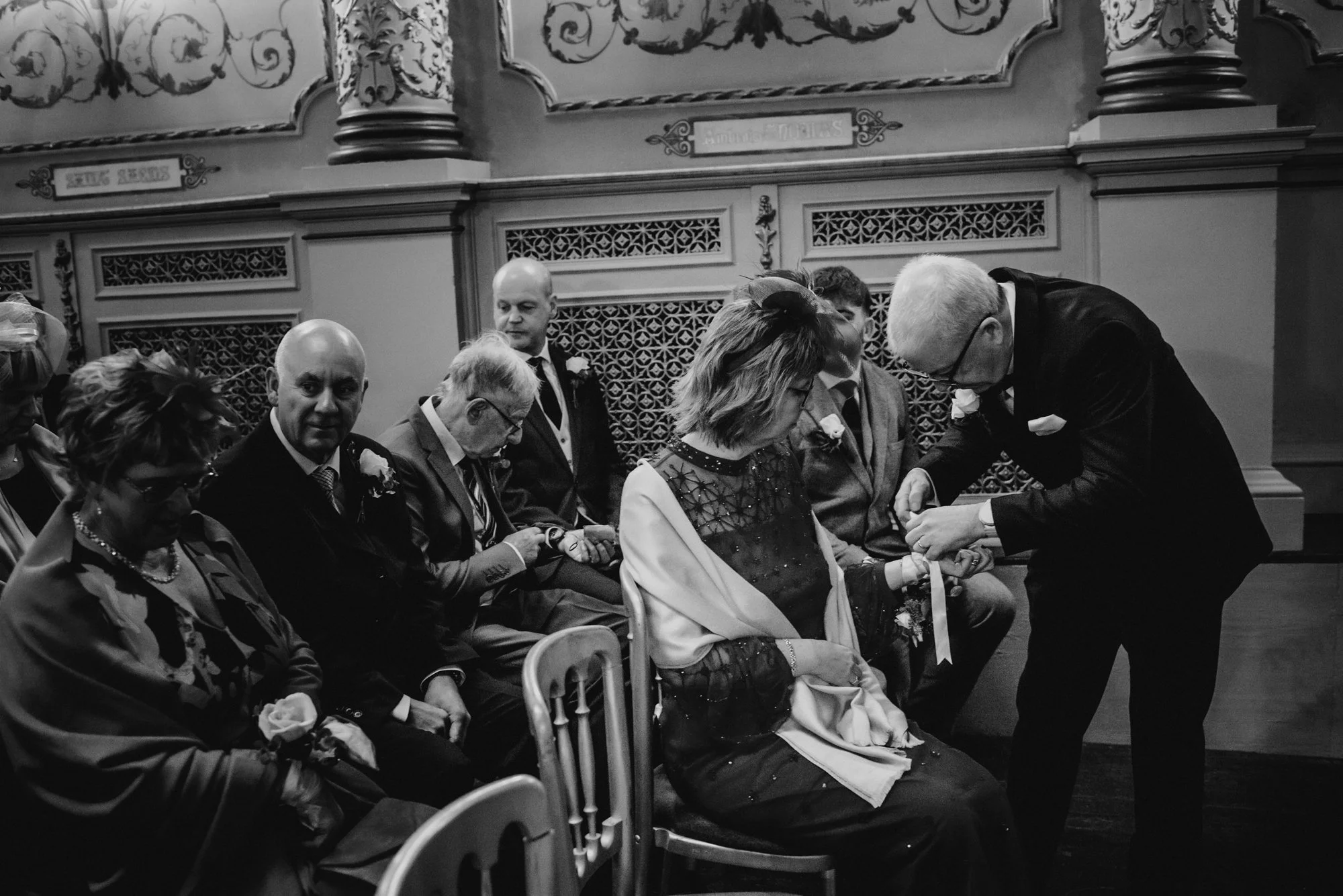 A black-and-white photo of a wedding ceremony inside a decorated church, with a man in a tuxedo placing a ring on a woman's finger. Several people are seated, watching the ceremony, and are dressed formally.