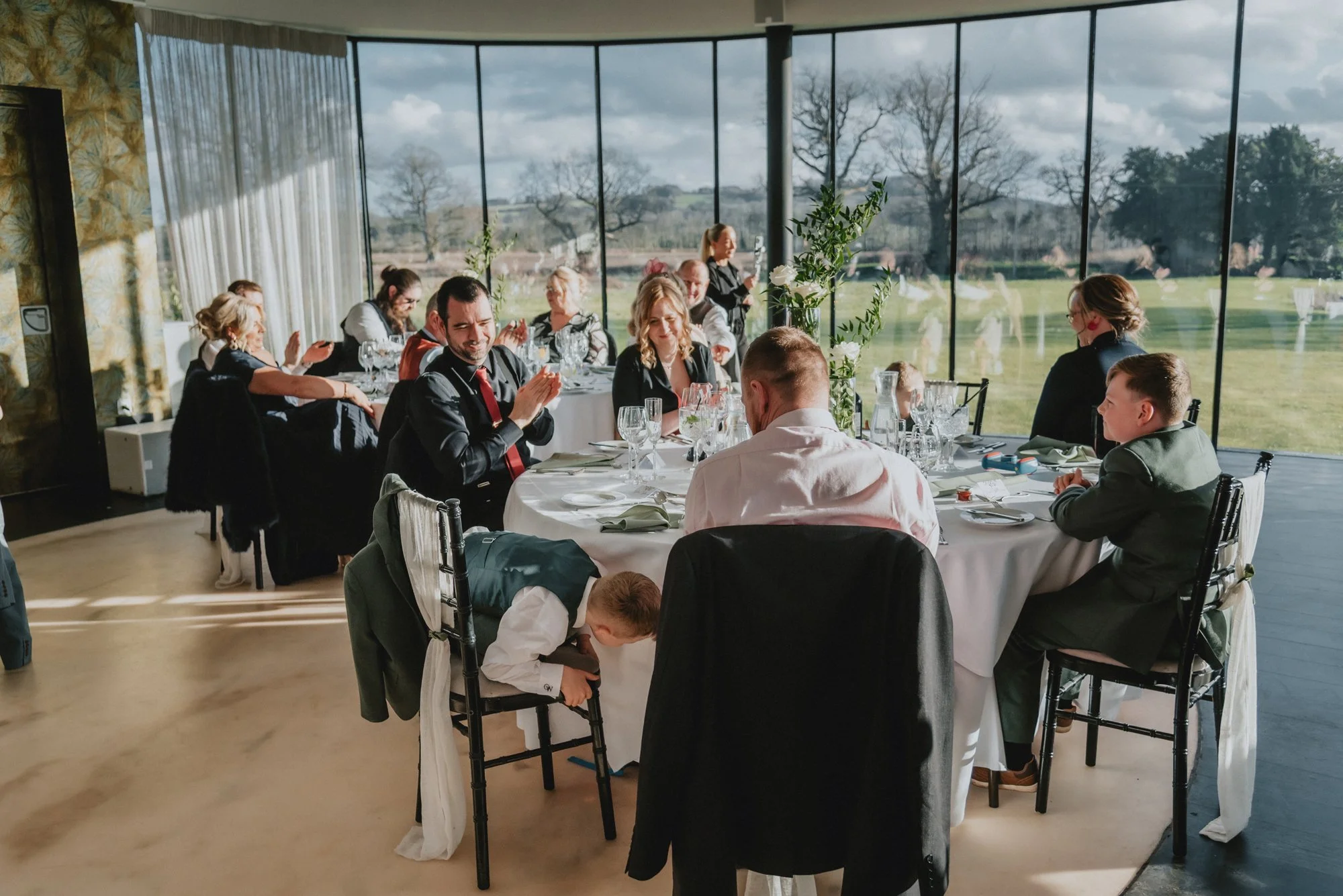 People attending a formal event or celebration at round tables inside a venue with floor-to-ceiling windows showing an outdoor landscape of trees and fields.