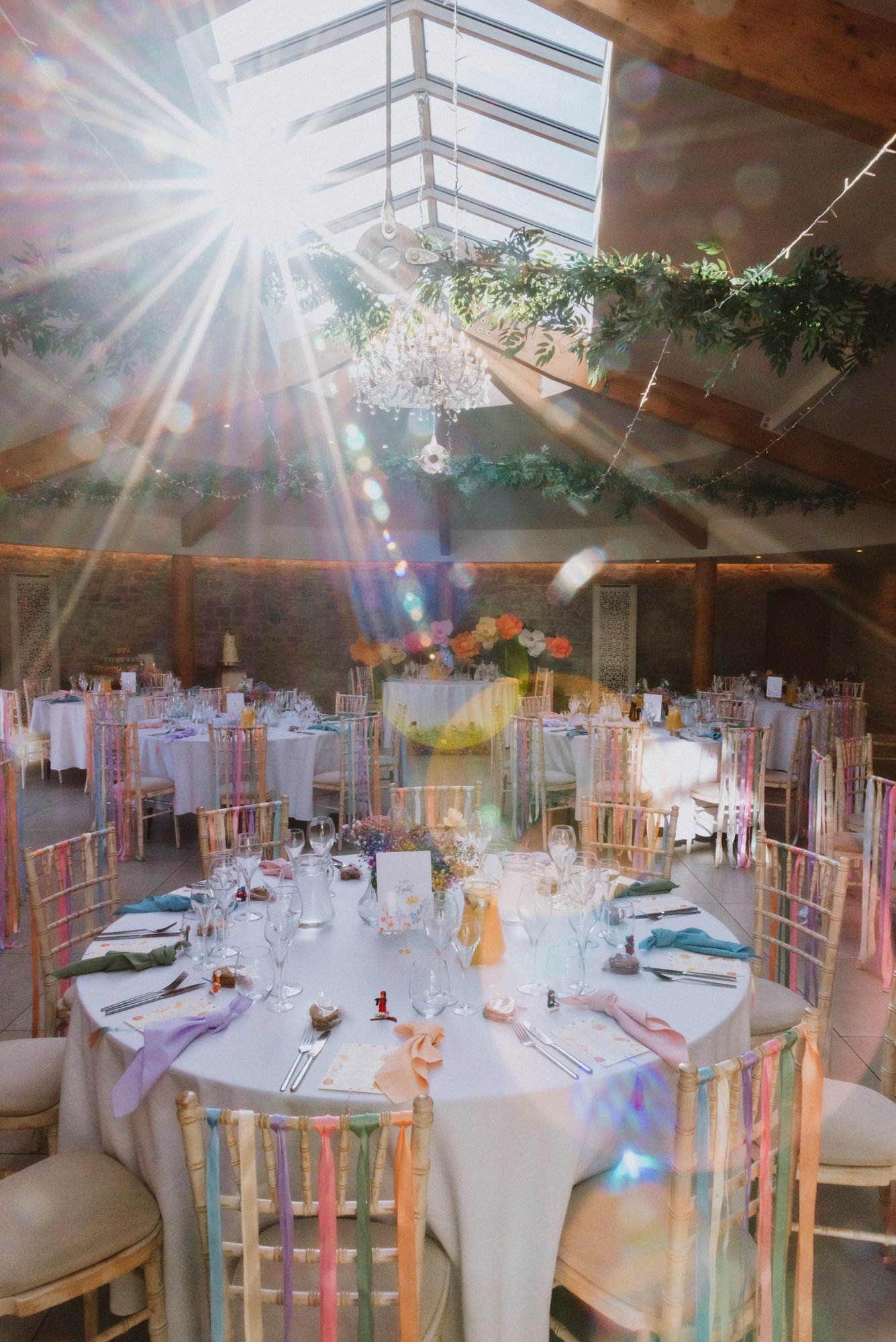 A decorated indoor event space with sun rays coming through a skylight, featuring a round table with white tablecloths, pastel-colored napkins, flower arrangements, and set for a formal dinner, with additional tables and greenery hanging decorations.